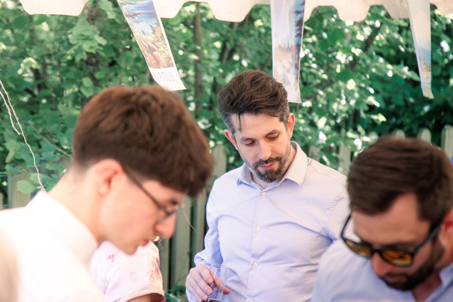 Three men gathered together looking down at something at an outdoor wedding celebration under canopy.