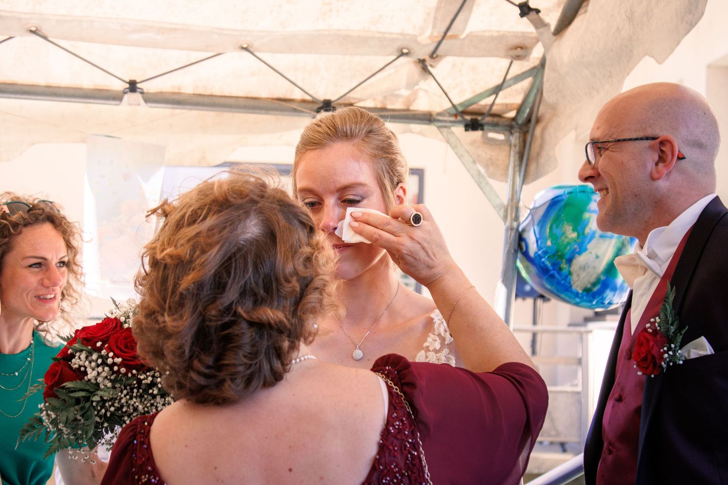 Emotional bride being comforted by guest while groom smiles at wedding reception under tent.