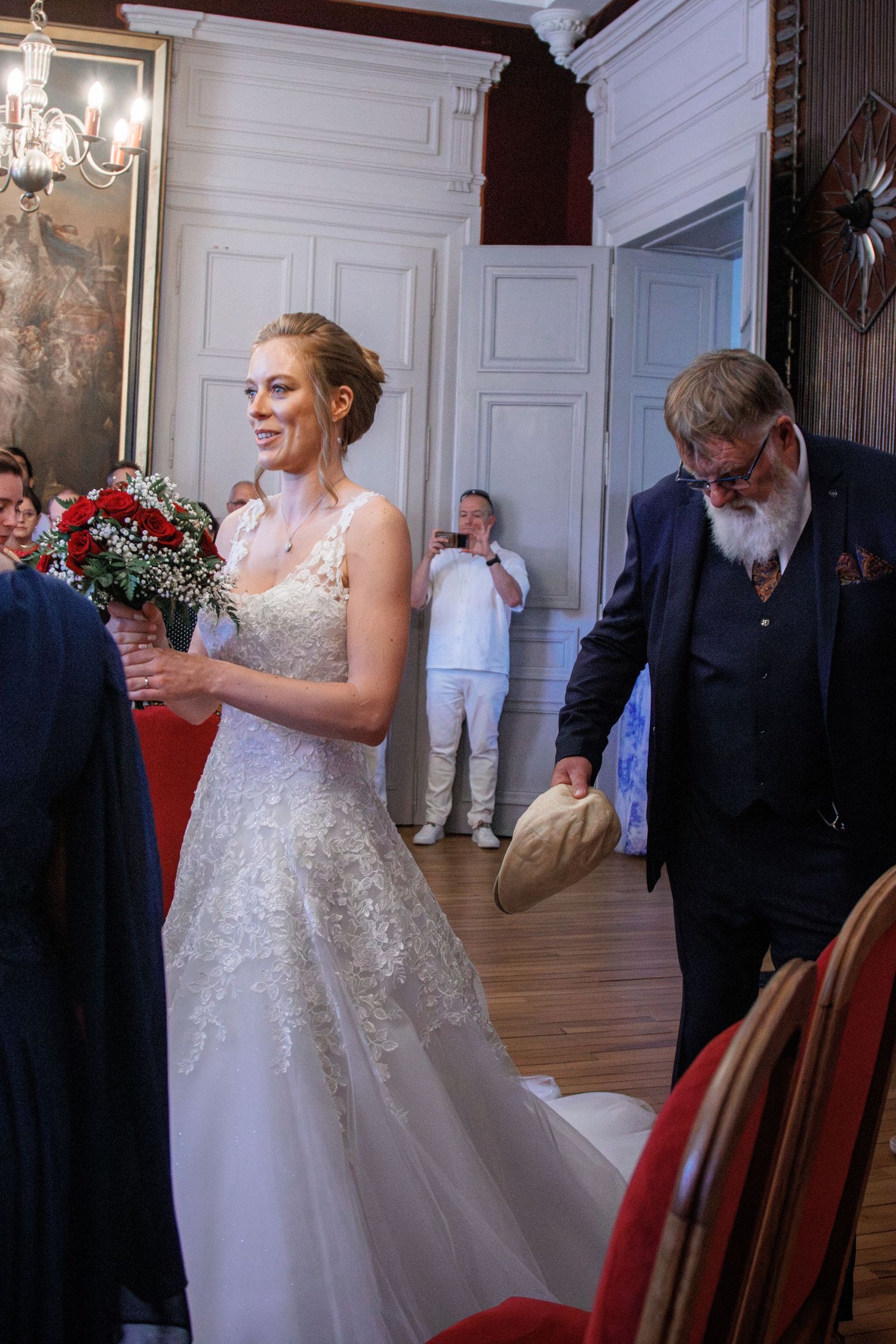 Bride in lace wedding gown holding red rose bouquet walks with bearded man in navy suit during indoor wedding ceremony.