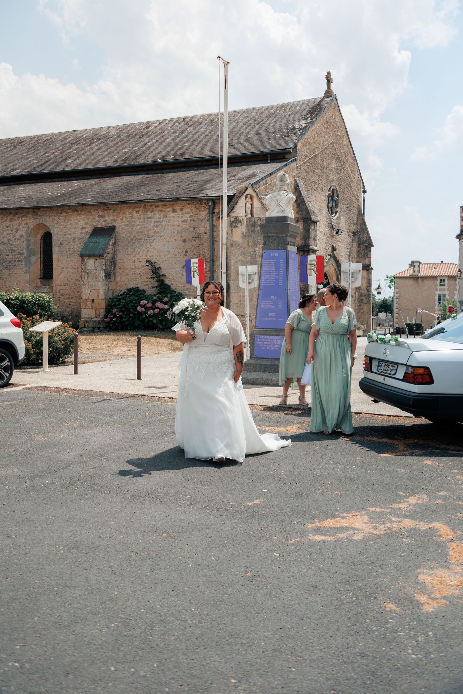 Bride in white gown walks with bridesmaids in sage green dresses outside a stone church on a sunny wedding day.