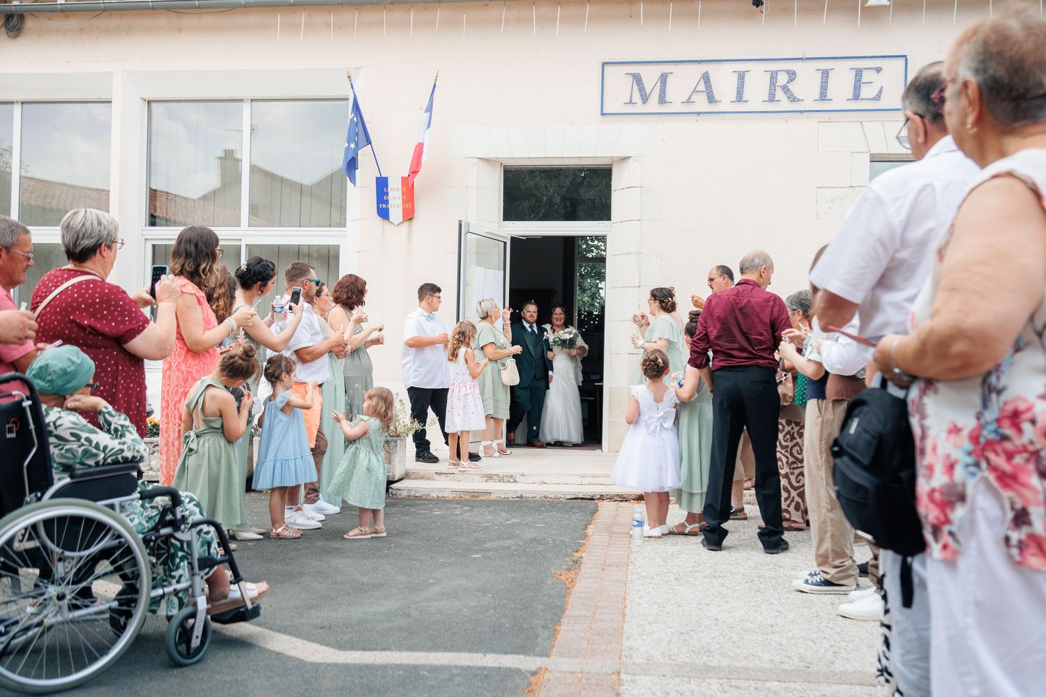 Newlyweds exit French mairie greeted by cheering guests and flower girls in blue dresses.