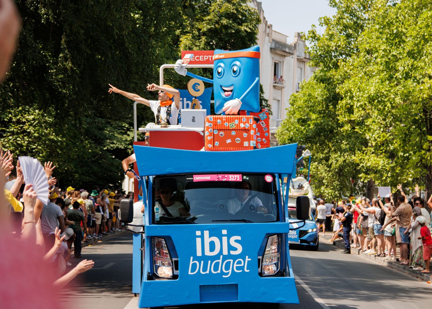 A bright blue Ibis Budget promotional vehicle with decorative elements participates in a street parade.