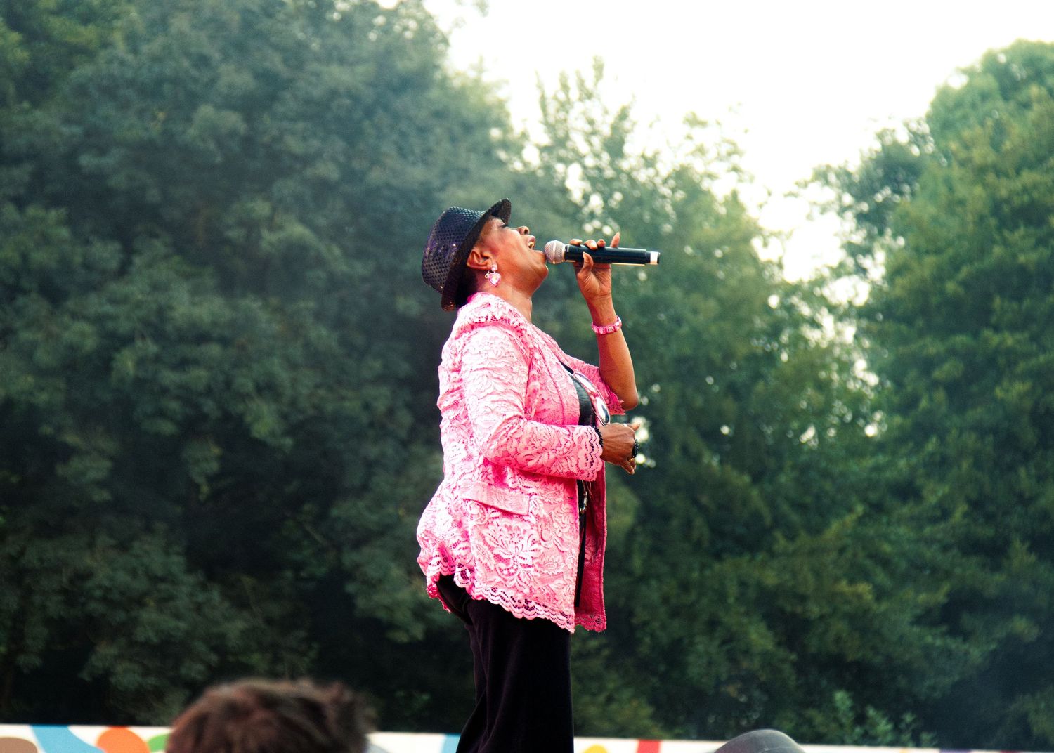 A performer in a pink top sings into a microphone while standing on stage during an outdoor concert.