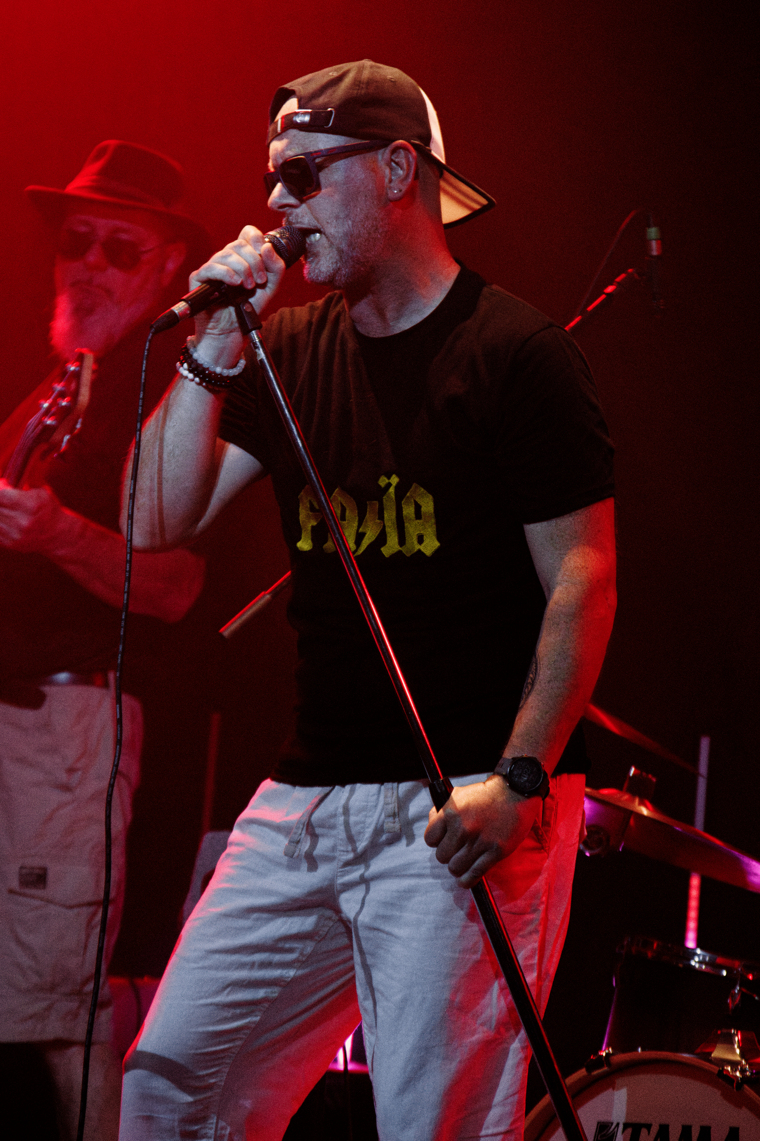 A performer in a black t-shirt and backwards cap sings passionately on stage under red lighting during a concert.