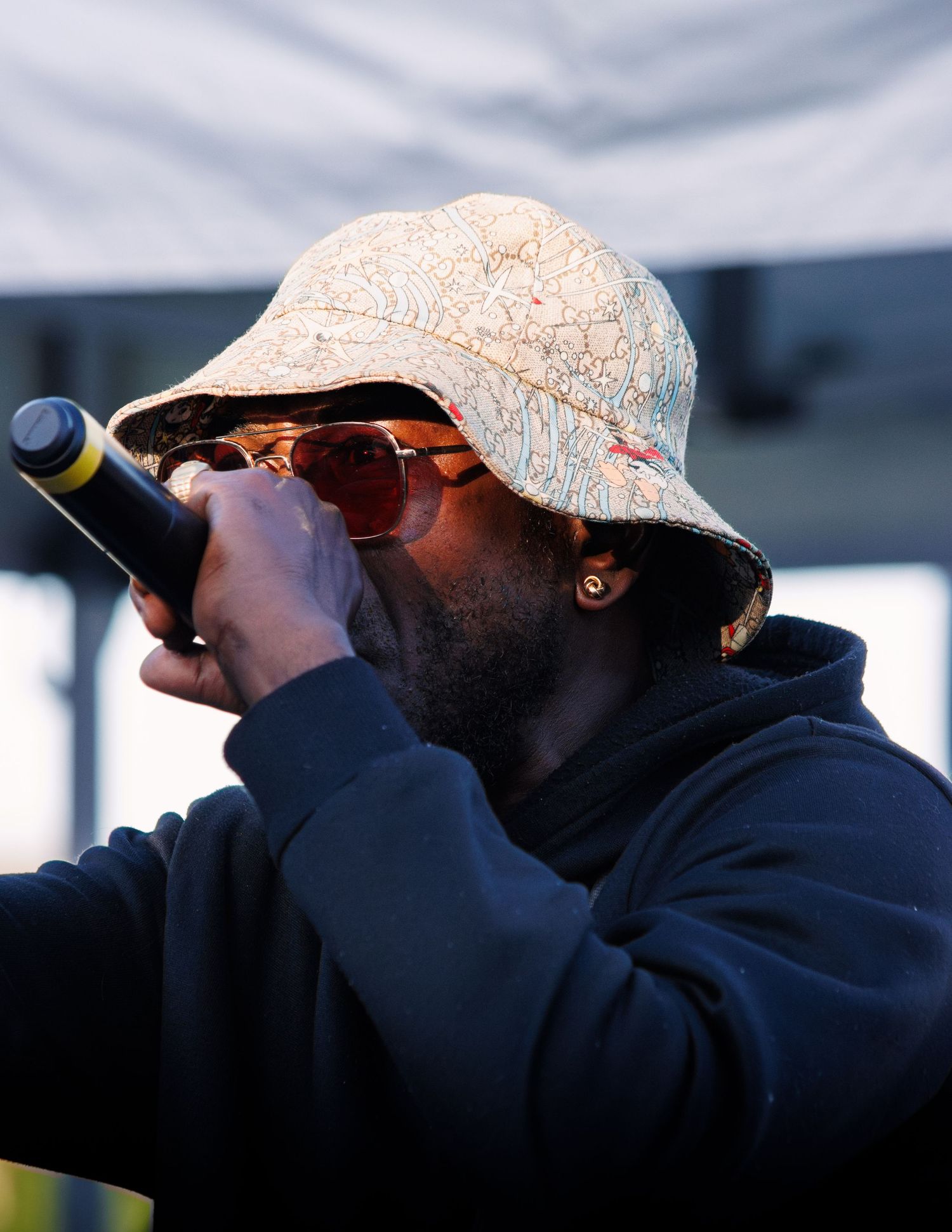 Performer wearing white bucket hat and sunglasses singing into microphone during outdoor concert.
