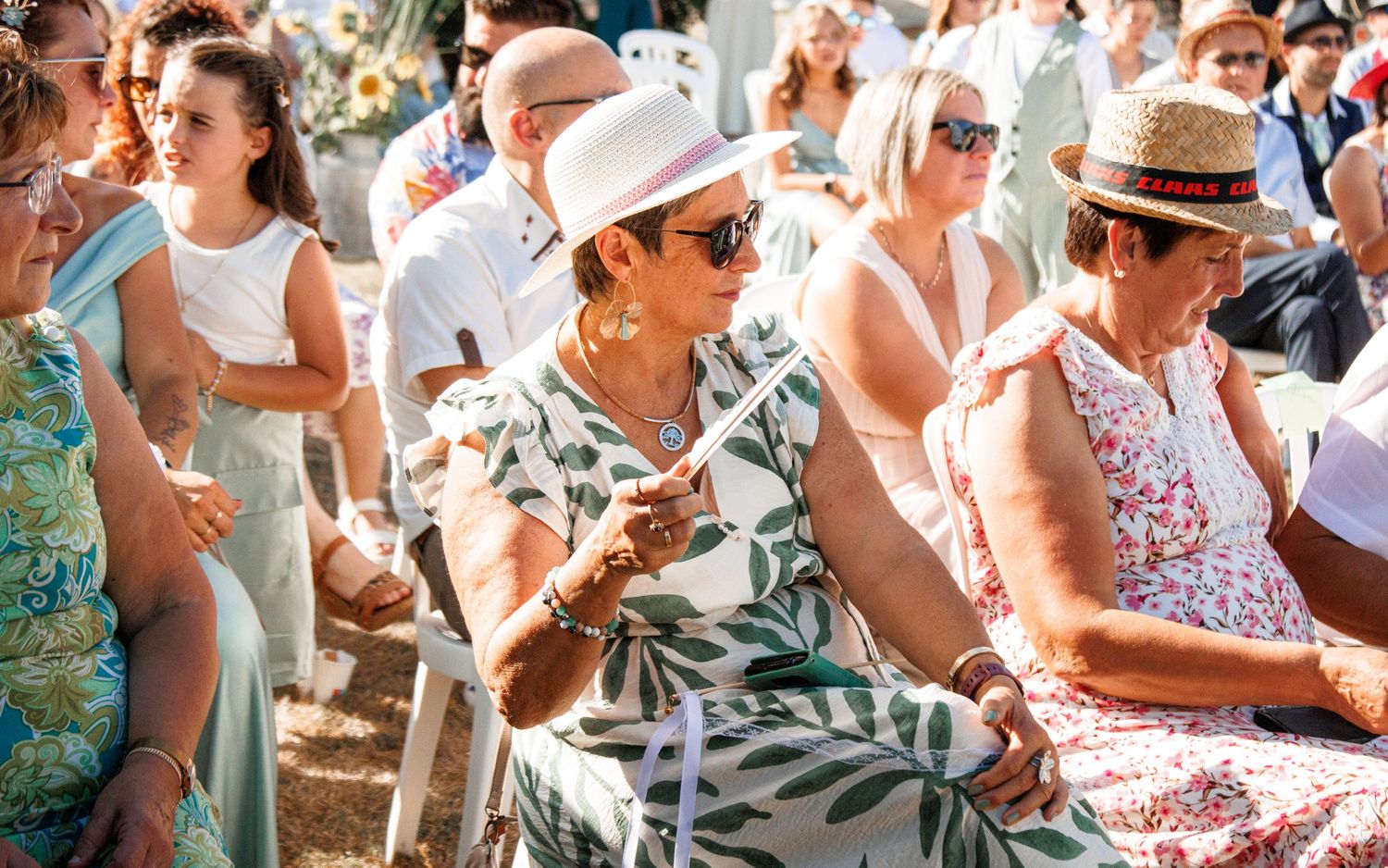 People gather outdoors at a sunny summer event wearing casual clothing and straw hats while enjoying refreshments.