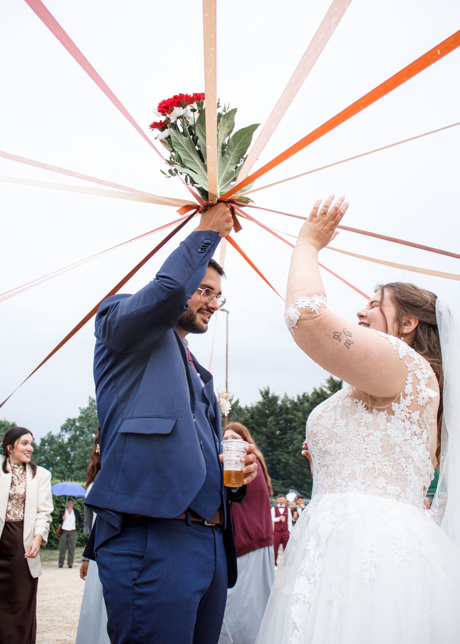 Bride and groom holding floral bouquet with colorful ribbons at outdoor wedding ceremony.