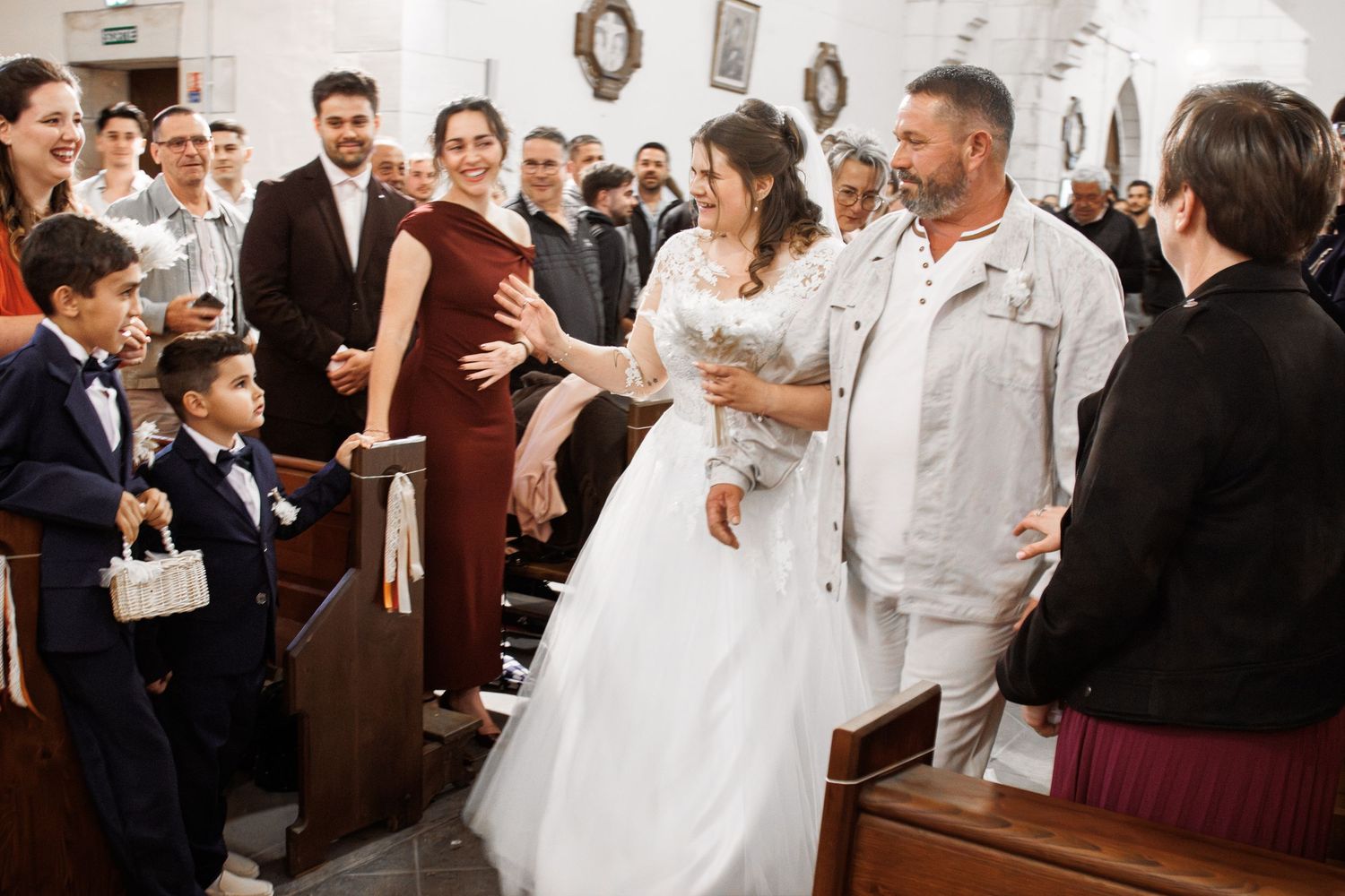 Bride in white gown greets guests in church aisle during wedding ceremony with smiling attendees in background.
