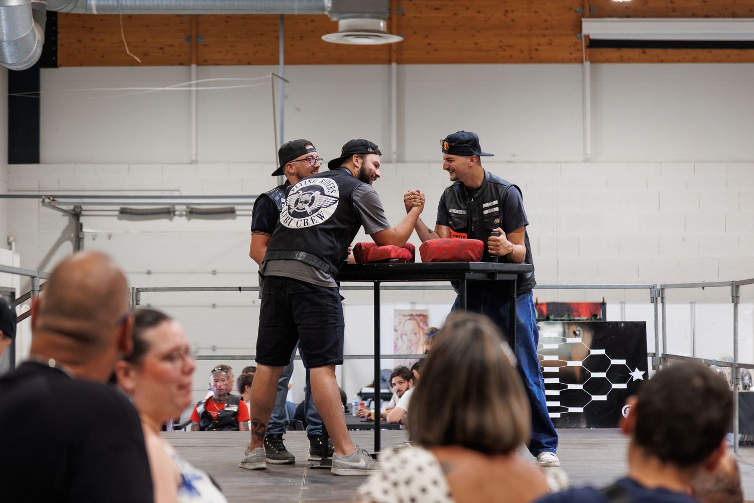 Two men arm wrestling on a red table at an indoor event with an audience watching in the foreground.