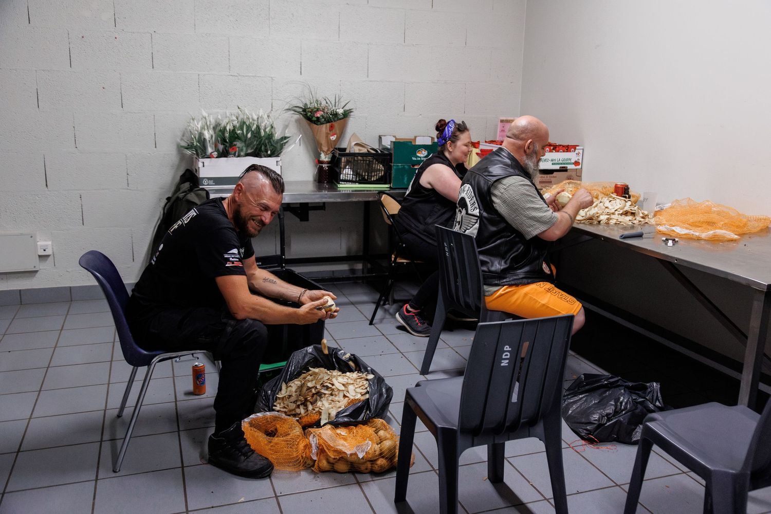 Three people sit in a small room preparing food packages, surrounded by bags and fresh flowers in the background.