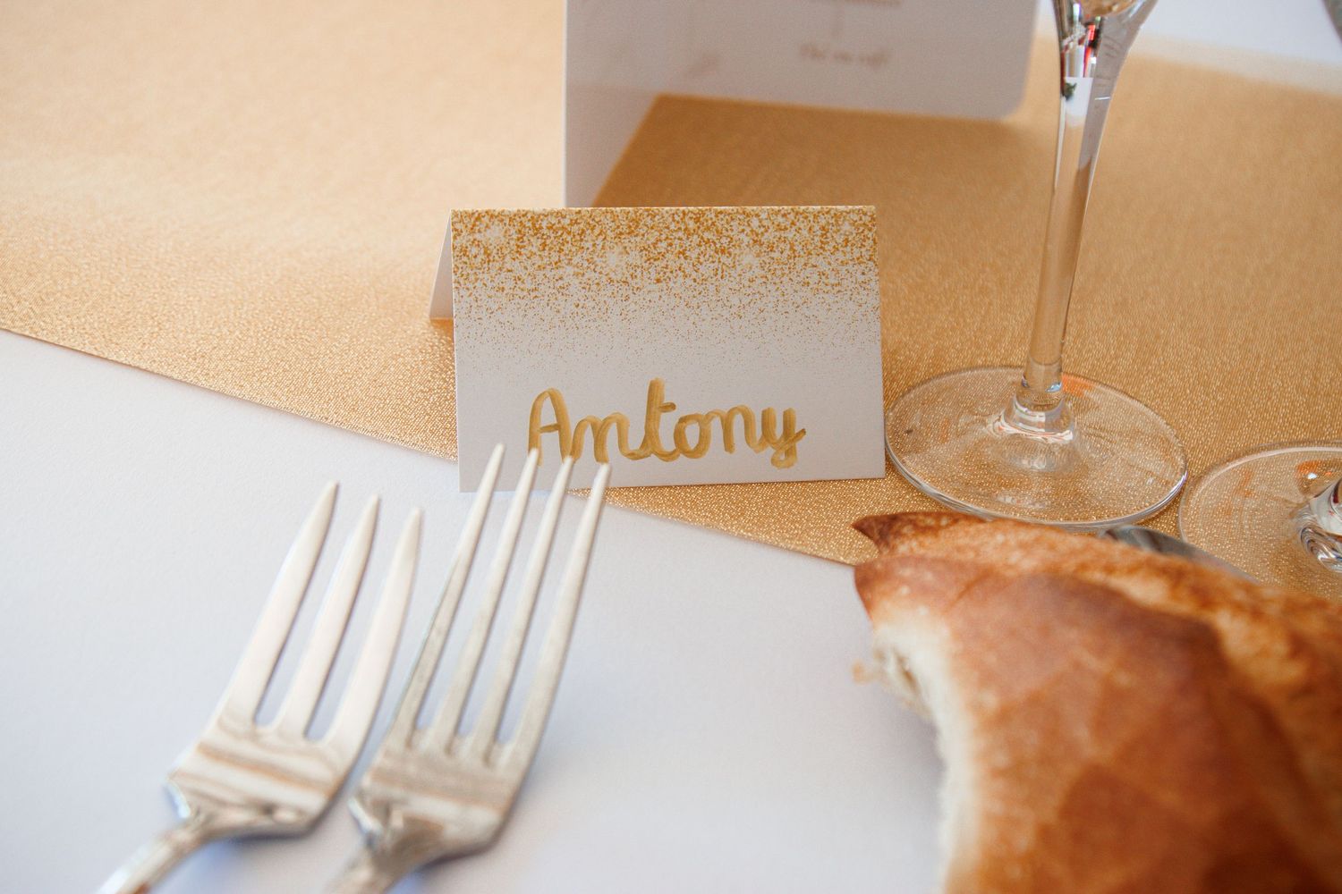 Wedding place card with name Antony in gold script beside two forks and bread on a table setting.