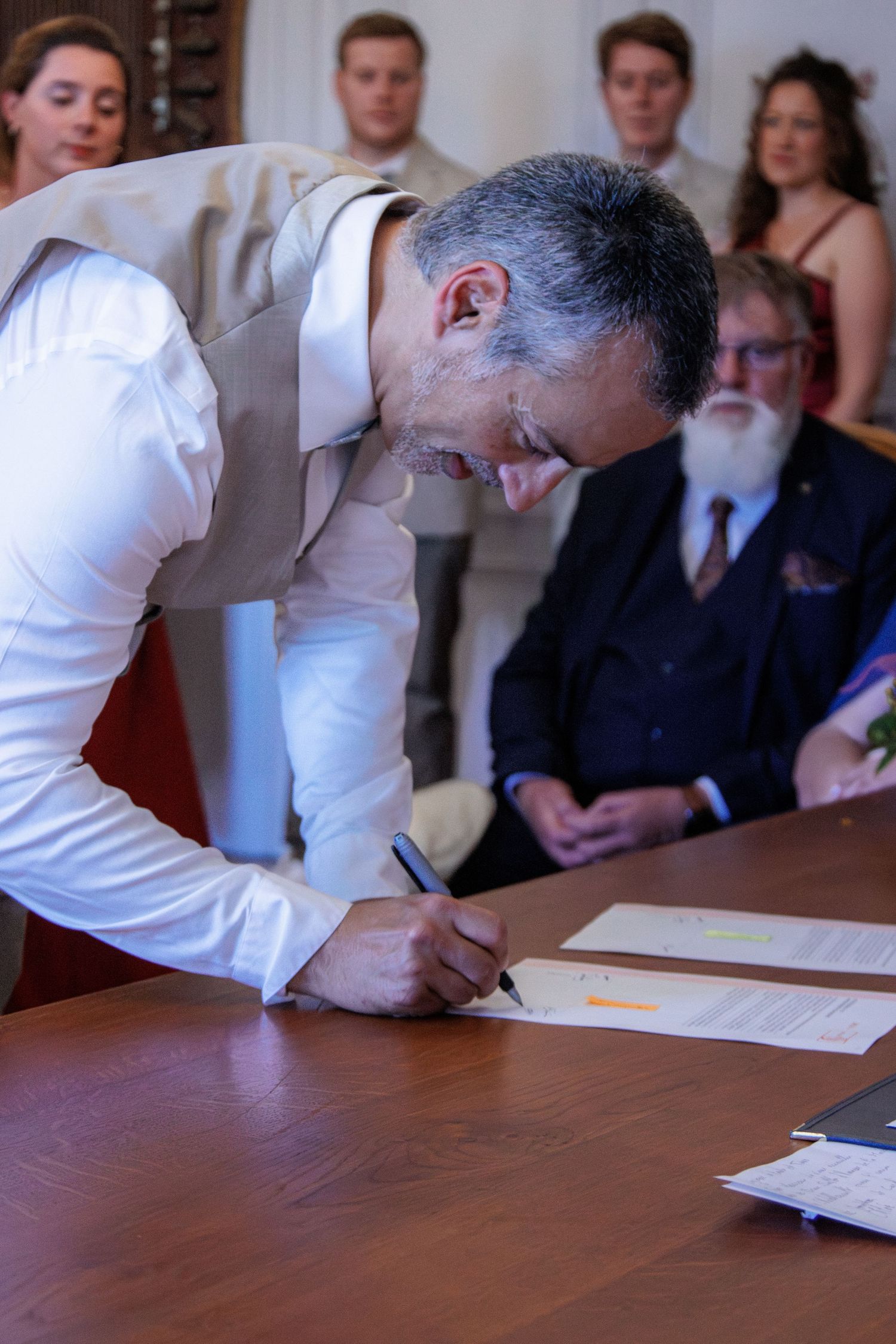Groom signing wedding documents at a wooden table during a civil ceremony with guests watching in the background.
