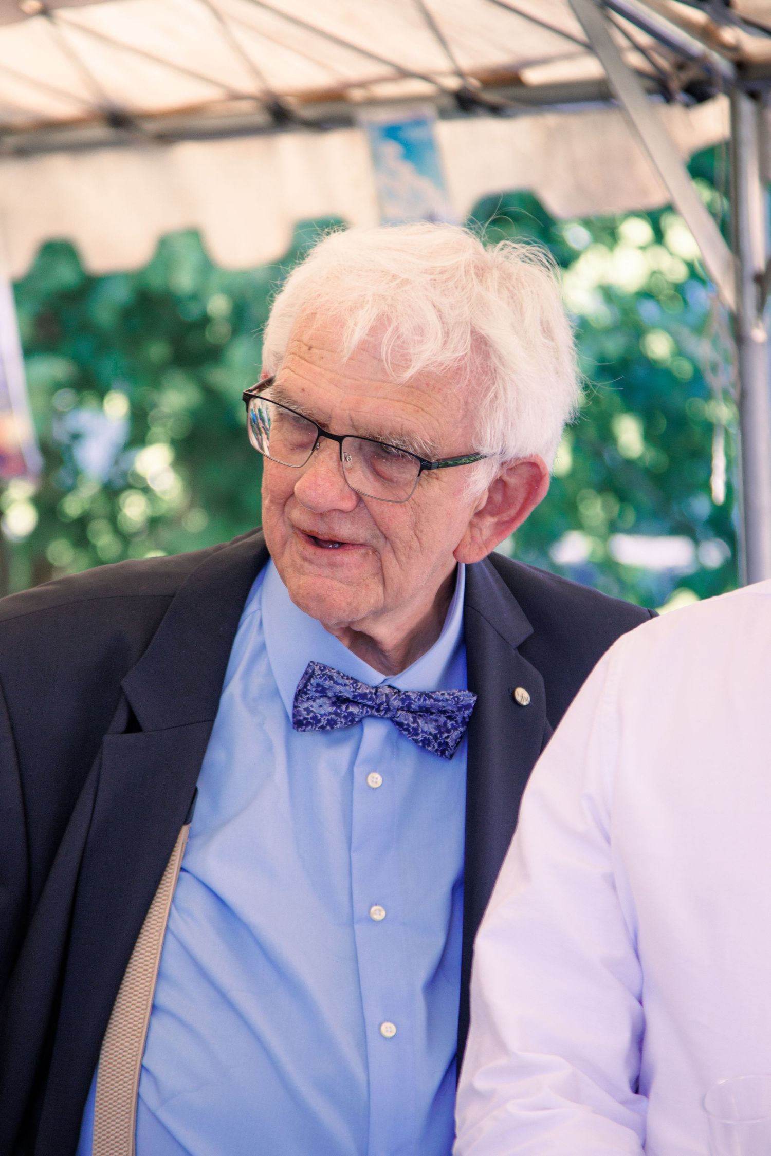 Elderly man with white hair and blue bow tie at an outdoor wedding reception.