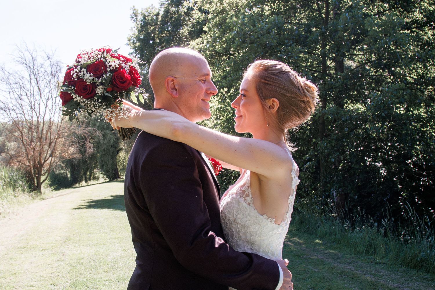 Bride and groom share a romantic embrace outdoors, bride holding a red bouquet in sunny garden.