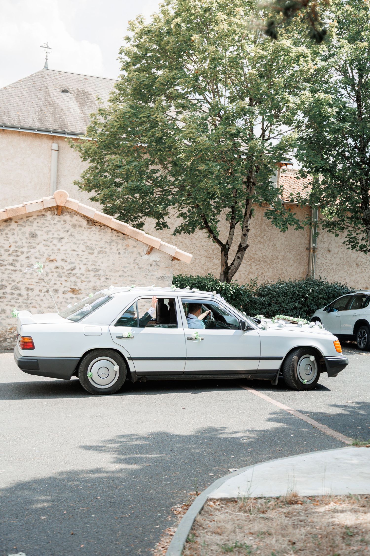 Classic white Mercedes wedding car decorated with white flowers parked outside stone church building.