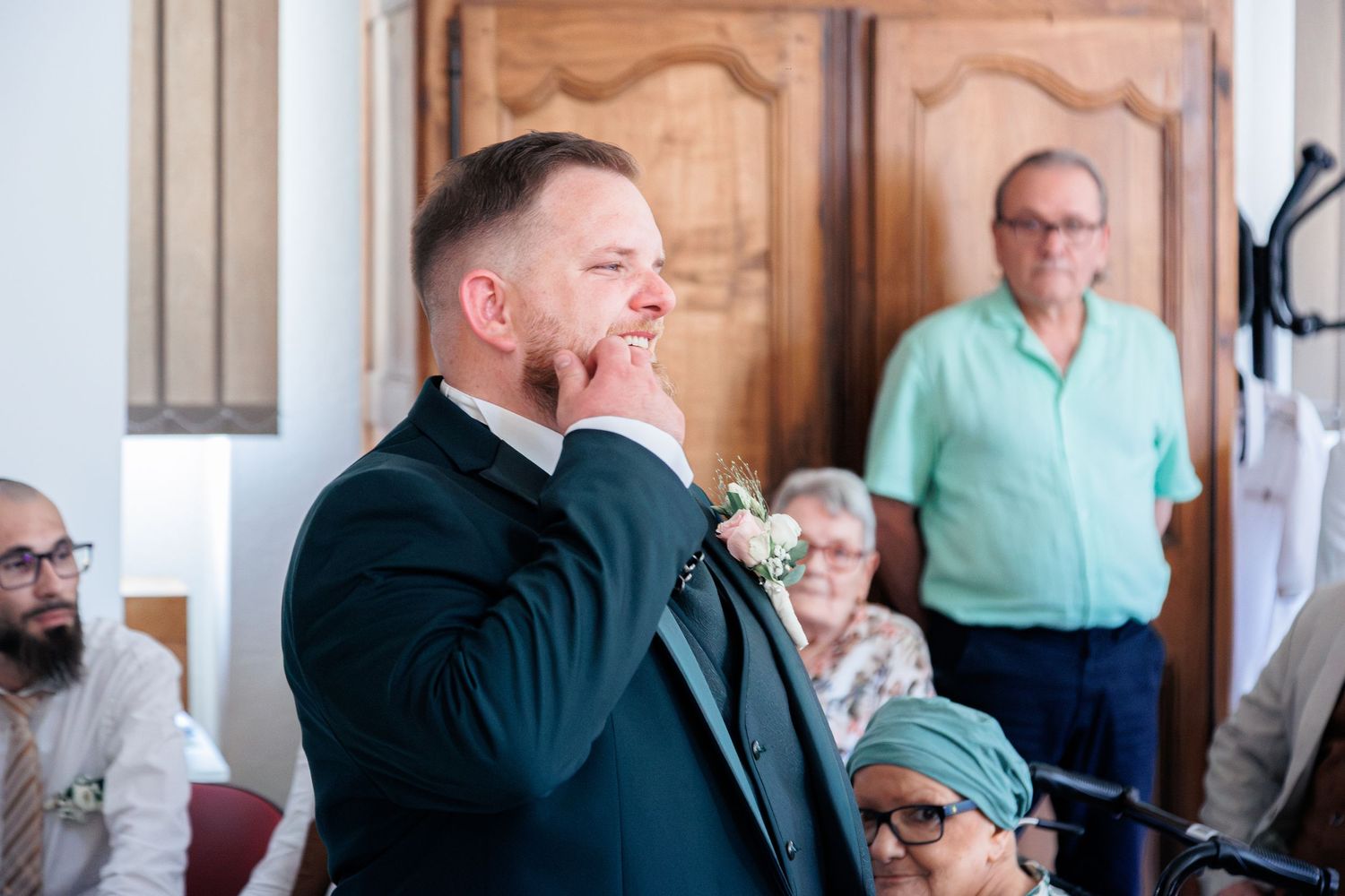 Groom in dark suit with boutonniere touches his chin thoughtfully during indoor wedding ceremony.