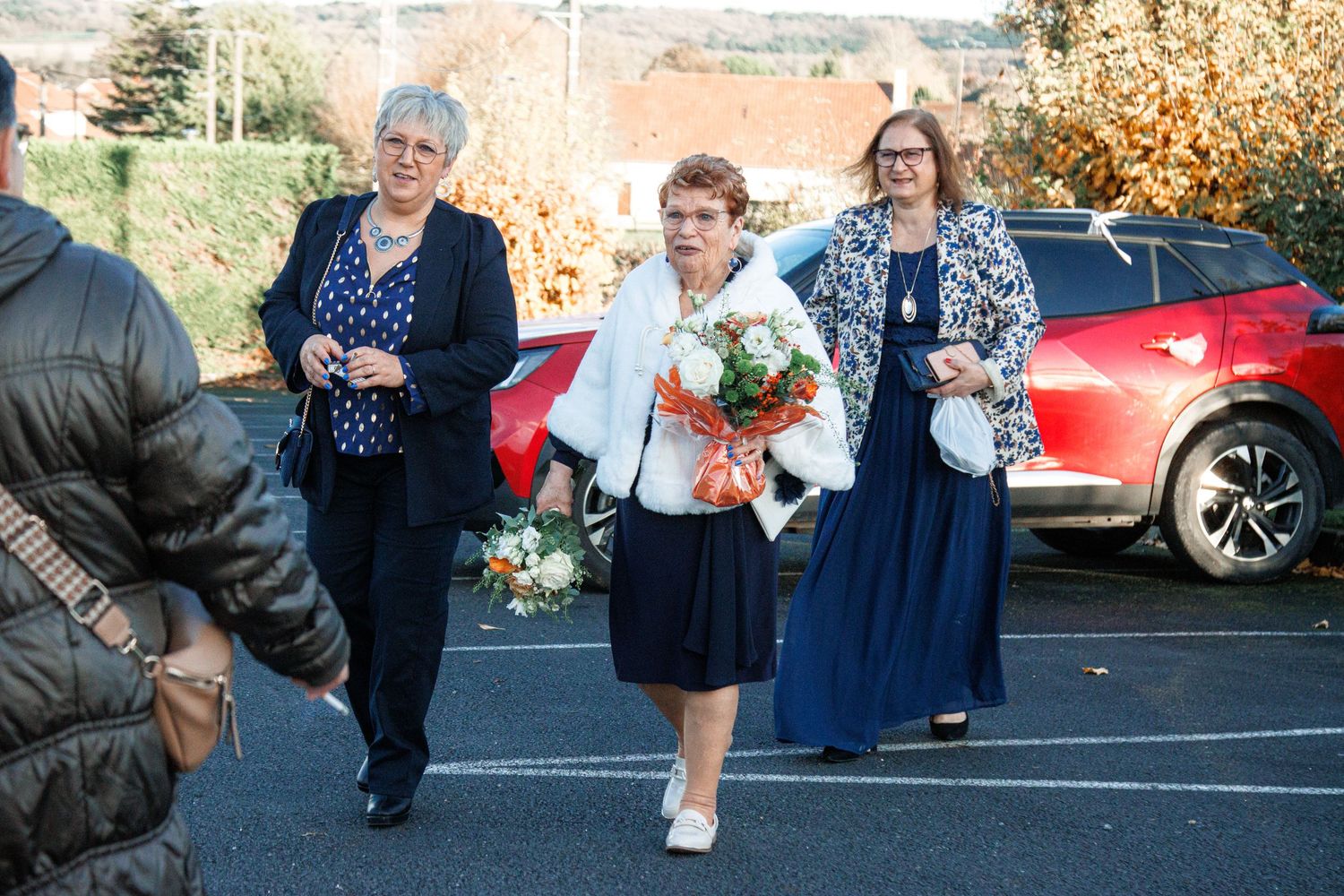 Three elegantly dressed women walking in a parking lot, carrying bouquets of flowers at a formal outdoor event.