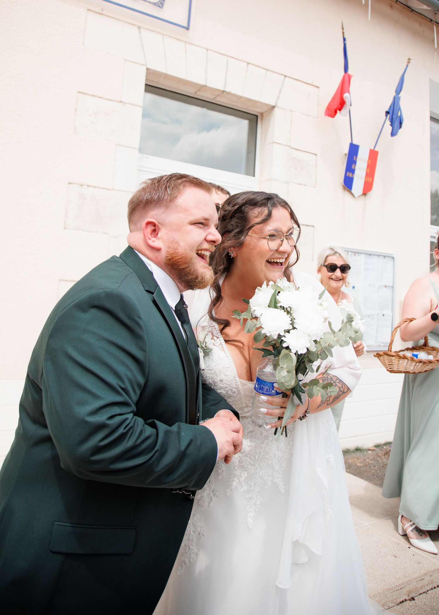A joyful bride and groom laugh outside a French building, she holds white flowers, he wears a green suit.