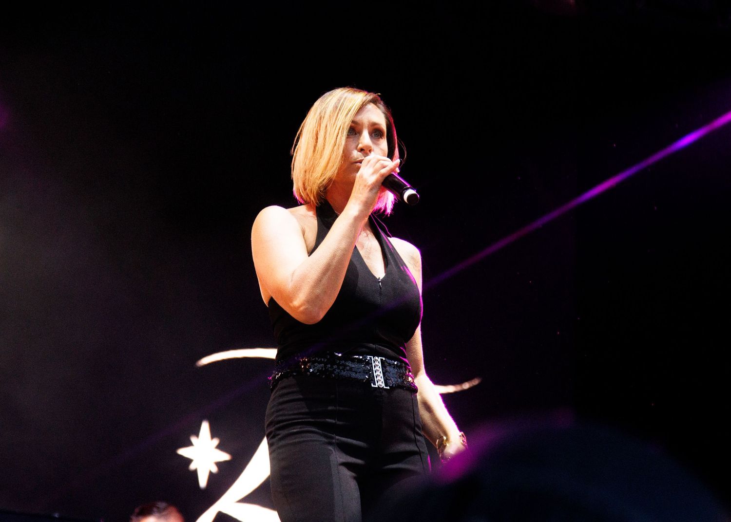 A concert performer in black attire sings into a microphone against a dark background with purple lighting.