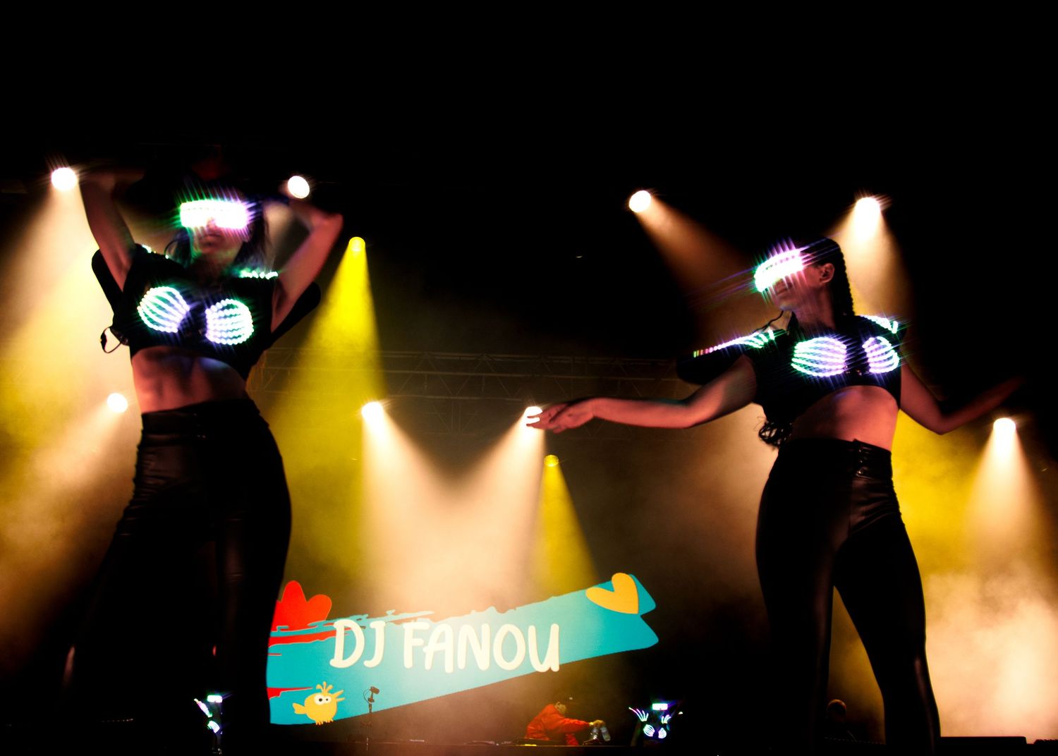 Performers in glowing LED costumes dance on a dark stage with yellow spotlights and DJ Panou text overlay.