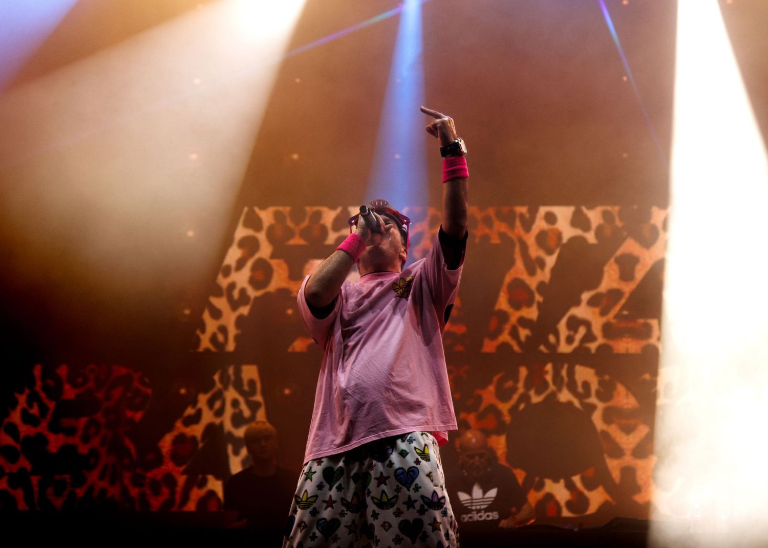 Performer on stage raises arm during concert with dramatic lighting and geometric pattern backdrop.