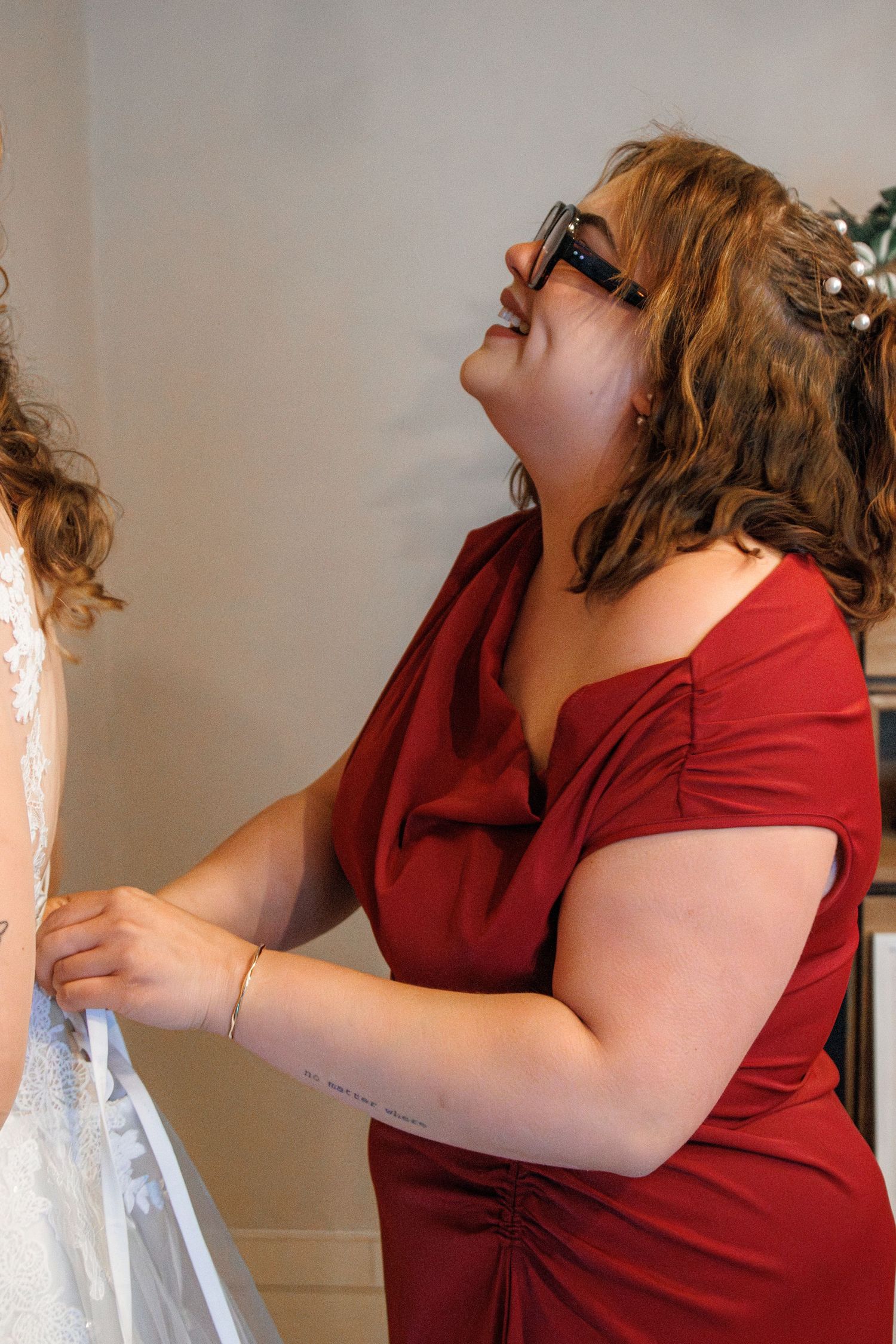 Bridesmaid in red off-shoulder dress helping bride with wedding gown buttons during bridal preparation.