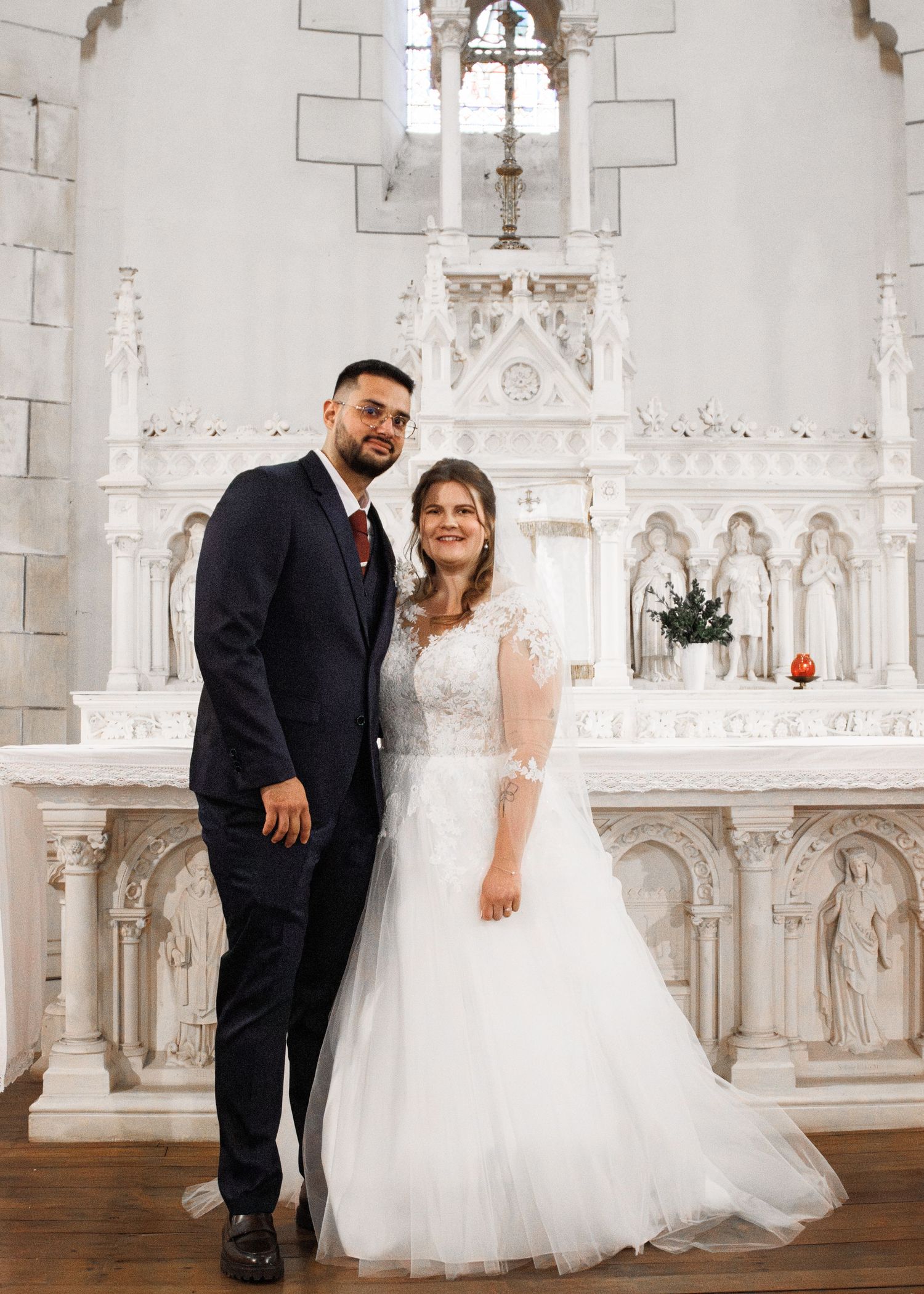 Bride in white lace gown and groom in navy suit pose together at ornate white church altar on wedding day.