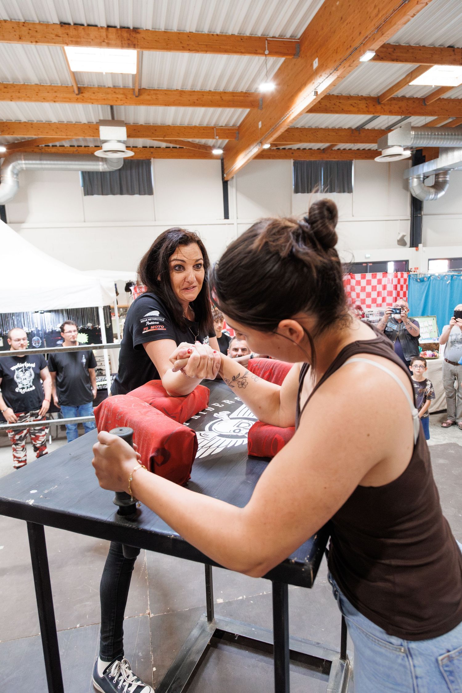 Two women competing in arm wrestling at an indoor tournament, with spectators watching in the background.