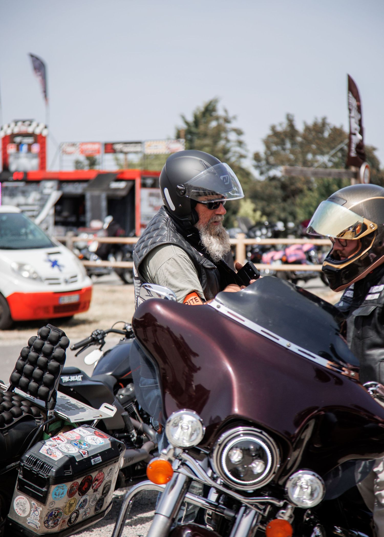 Bearded man wearing helmet and sunglasses riding a dark maroon Harley-Davidson motorcycle at an outdoor biker event.