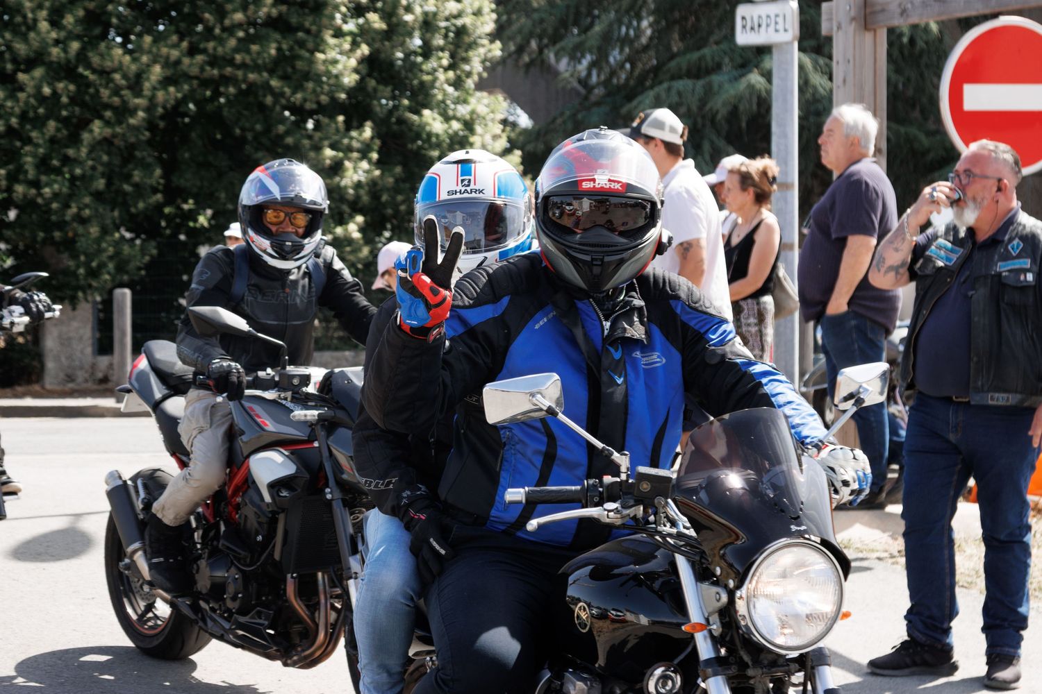 Motorcyclists in blue helmets and gear ride through a sunny street, with bystanders visible in the background.