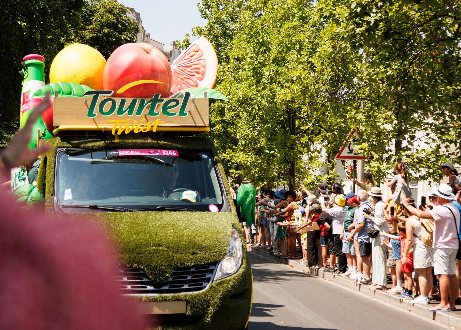 Tourrel branded promotional vehicle with giant fruit decorations drives through a crowd-lined street.