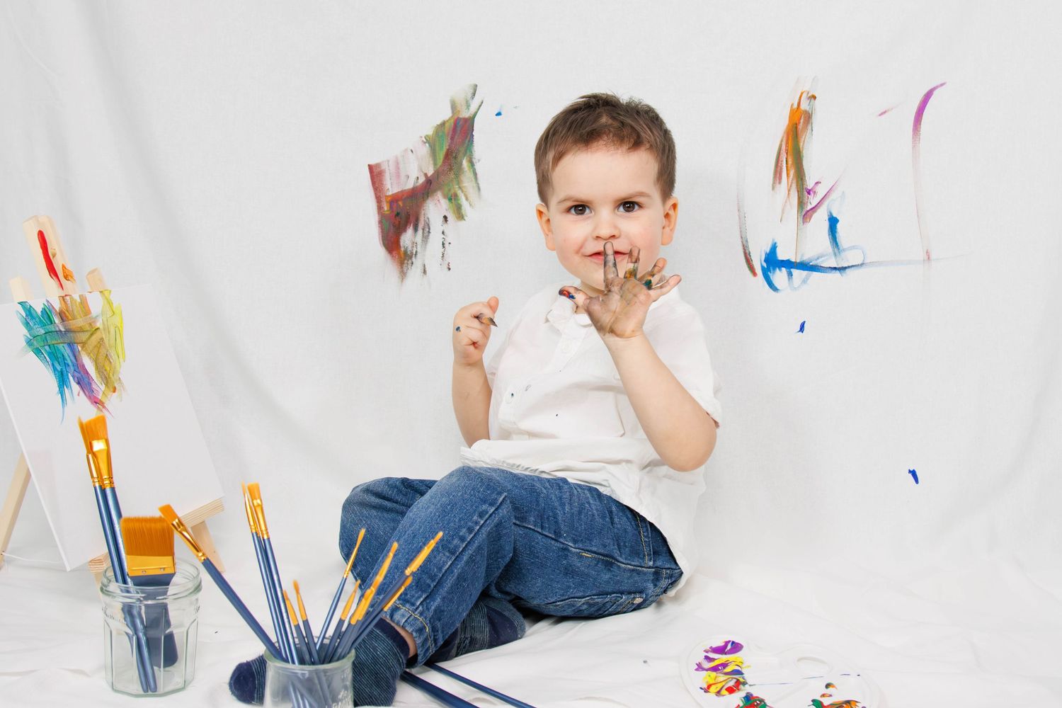 Child in white shirt sitting thoughtfully with art supplies and colorful wall paintings.