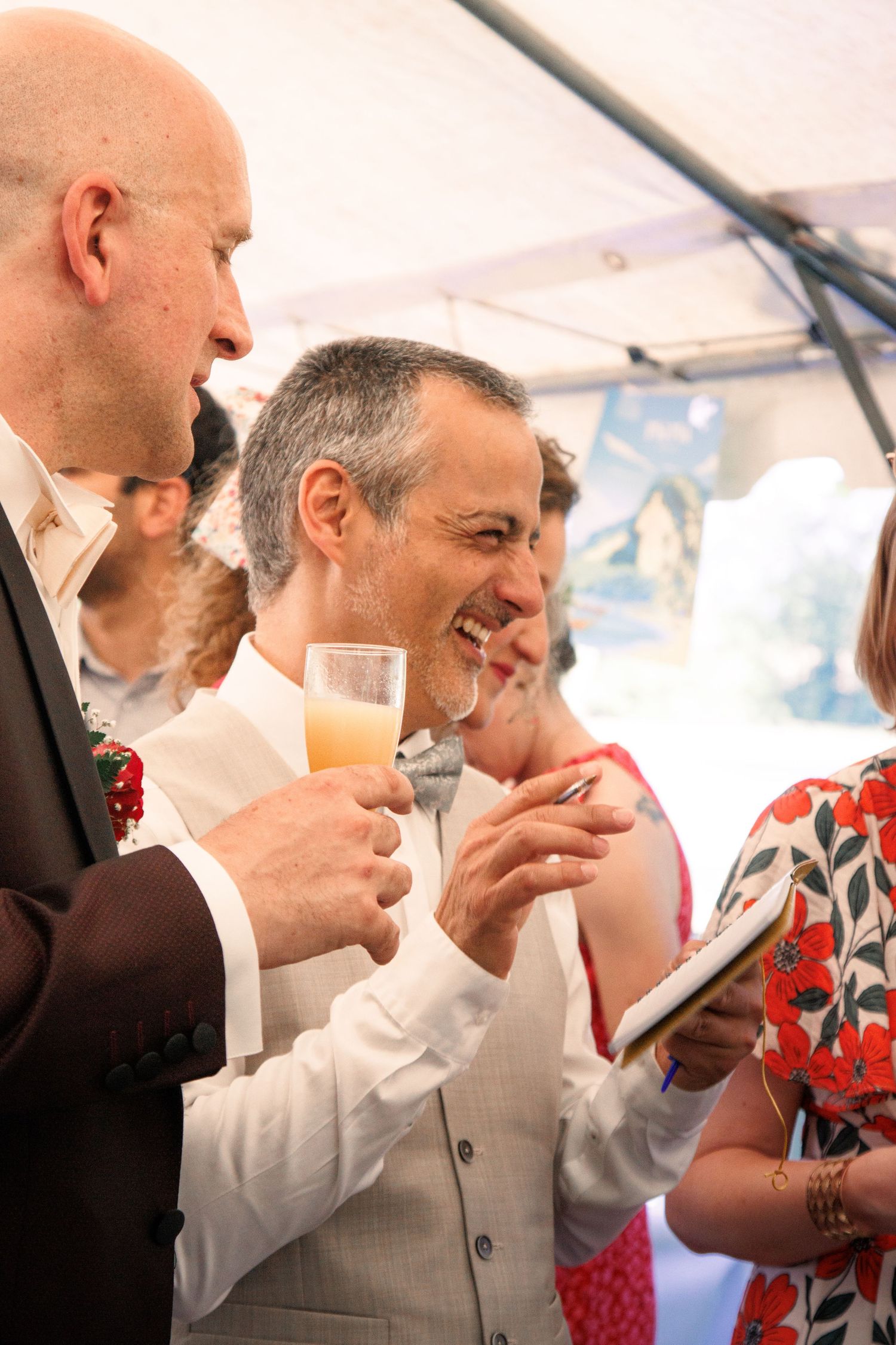Man in white shirt laughs joyfully while holding champagne glass at an elegant outdoor wedding reception.