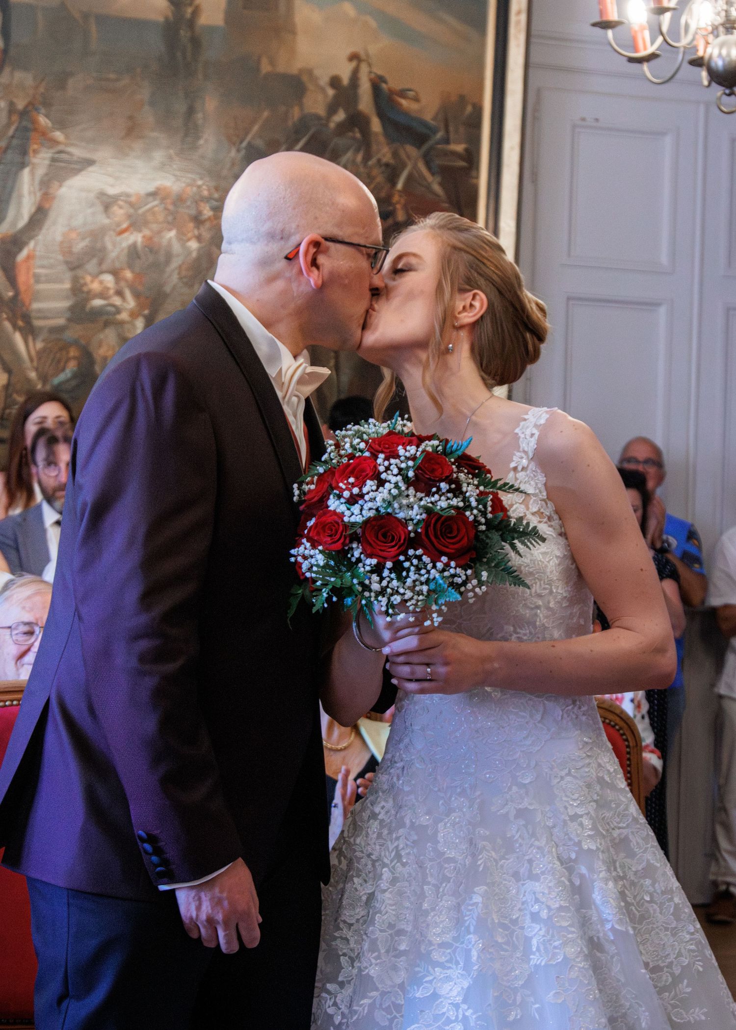 A groom in a purple suit kisses his bride holding red roses at their wedding ceremony.