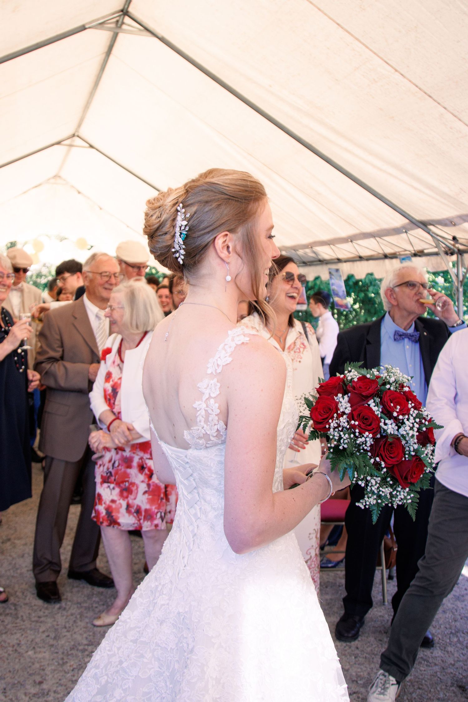 Bride in a lace wedding dress holding red roses bouquet, seen from behind, mingling with guests under a tent.