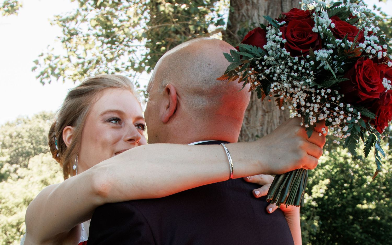Bride holding red roses bouquet embraces bald groom outdoors at wedding ceremony near green trees.