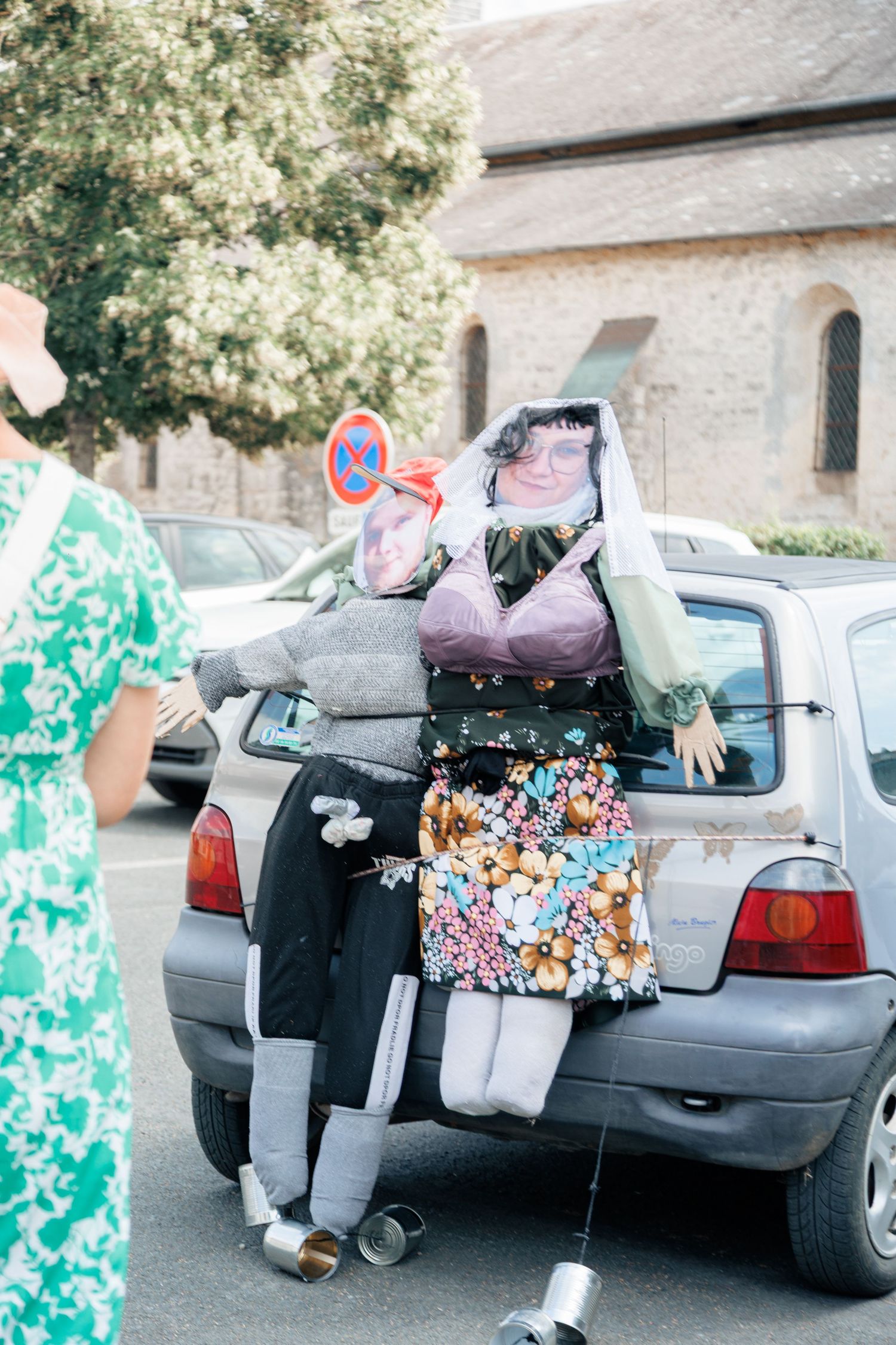 Humorous stuffed dummy bride decoration tied to car bumper as traditional French wedding prank outside church.