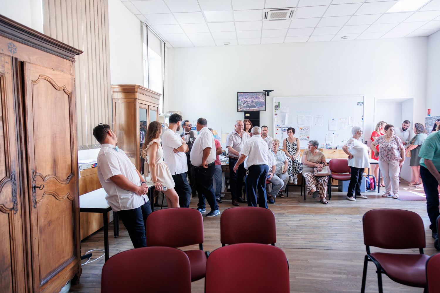 Wedding guests mingling in a reception hall with red chairs and wooden armoire during a celebration.