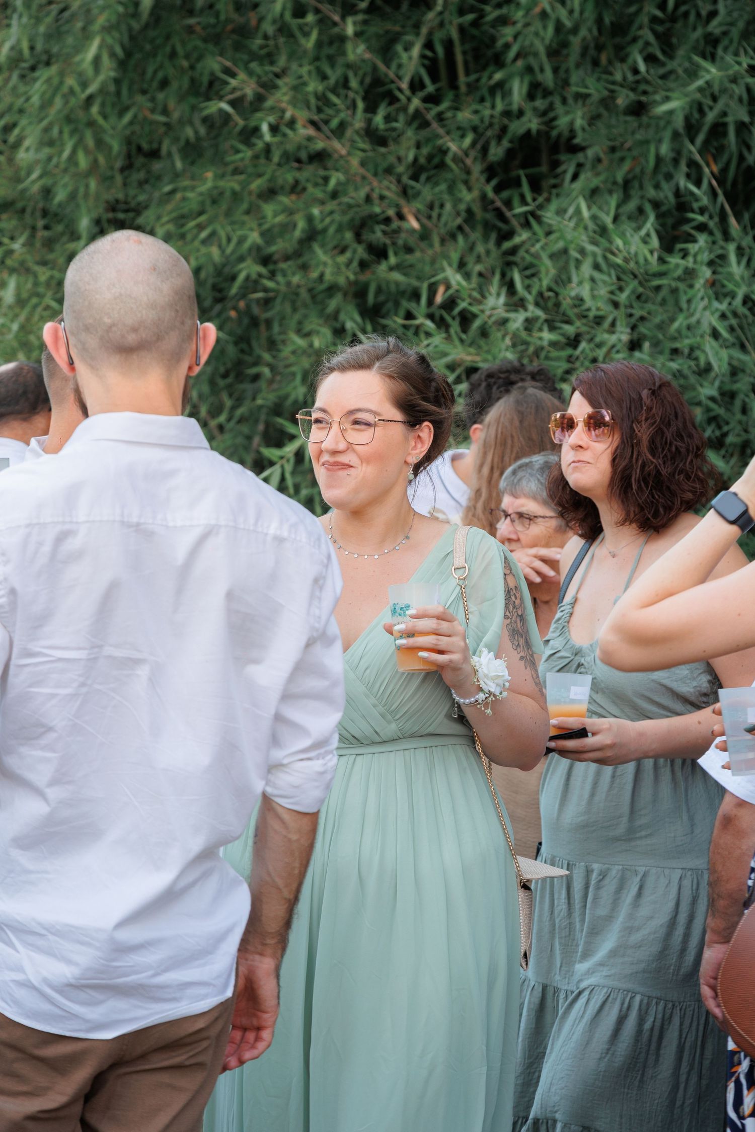 Wedding guests in sage green dresses socializing outdoors during cocktail hour with drinks in hand.