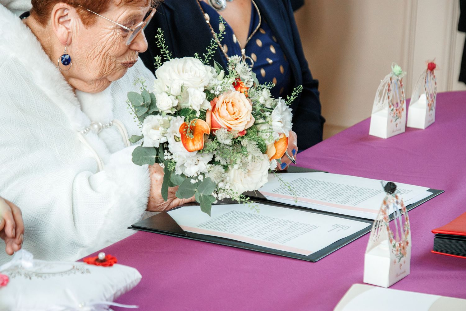 Elderly bride signing wedding documents at a purple table with a white and peach rose bouquet.