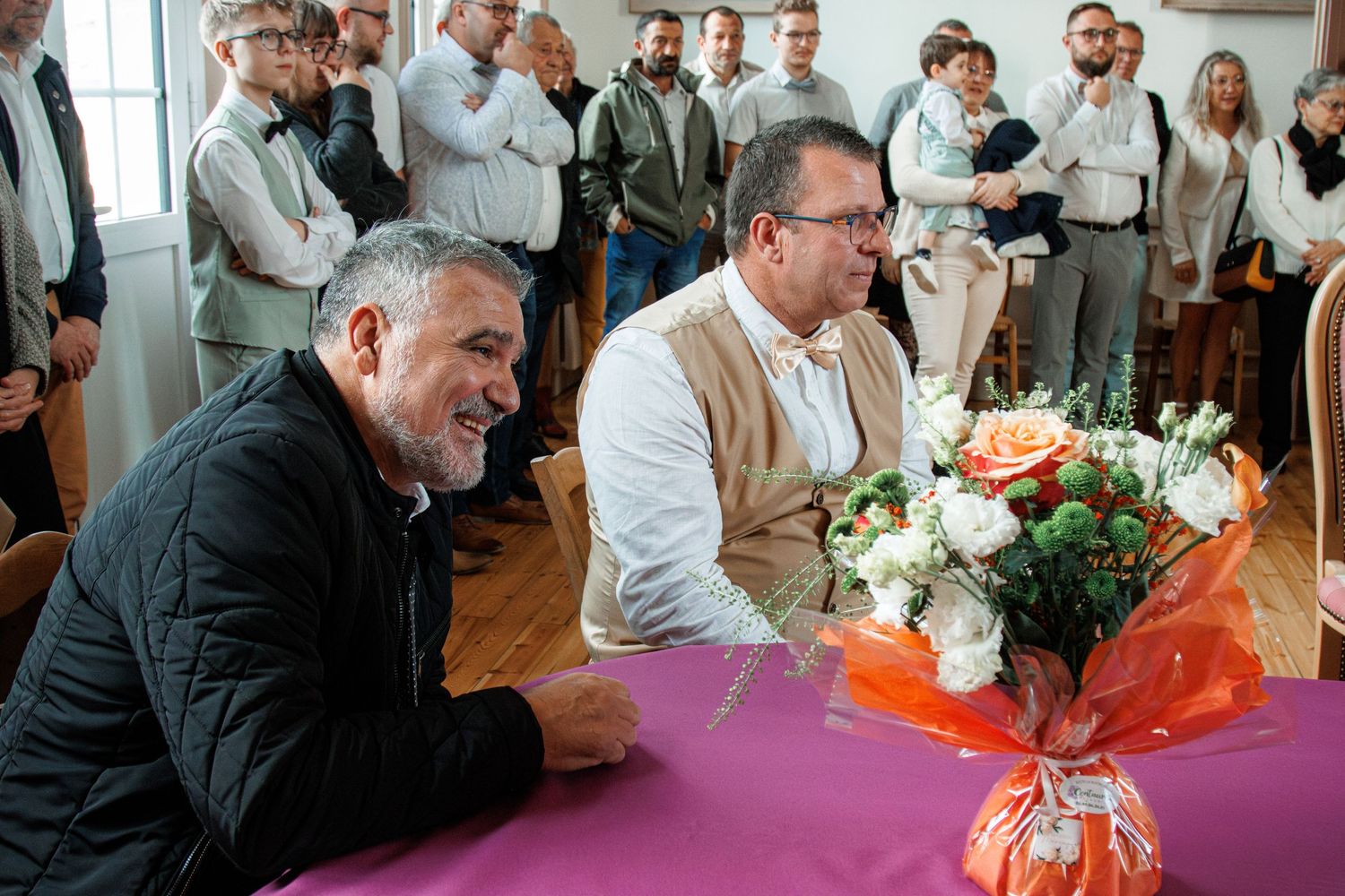 Two men sit at a purple-draped table with a floral bouquet, surrounded by guests at a formal indoor gathering.