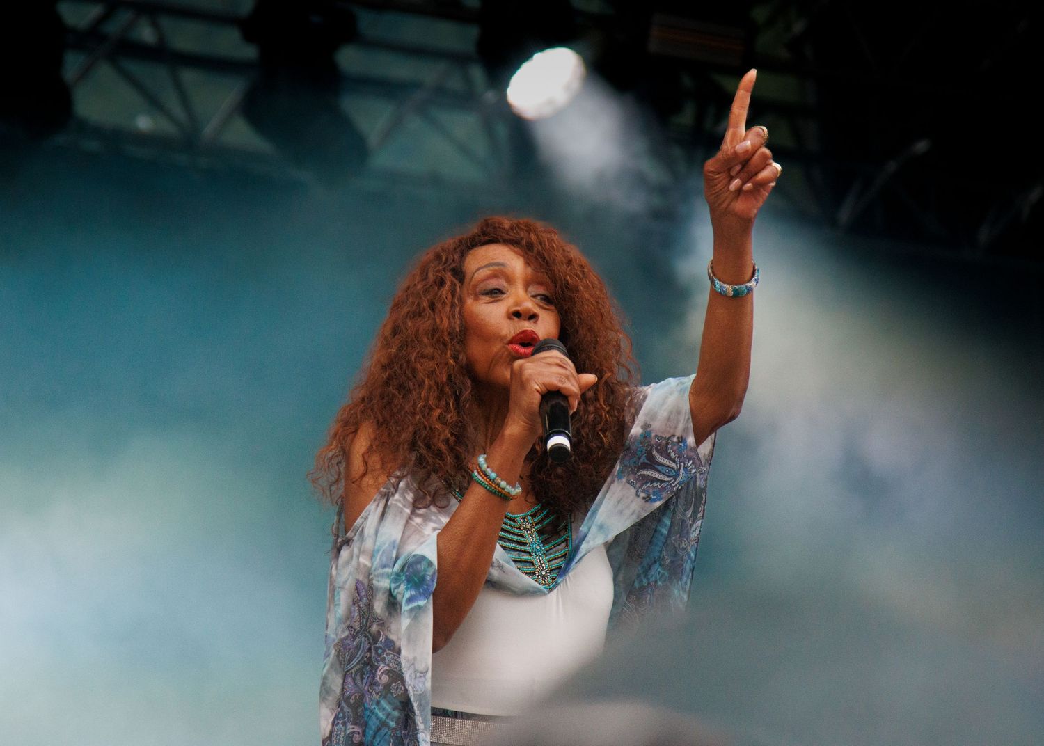 Singer with long curly red hair performs on stage, pointing upward under blue theatrical lighting and fog.