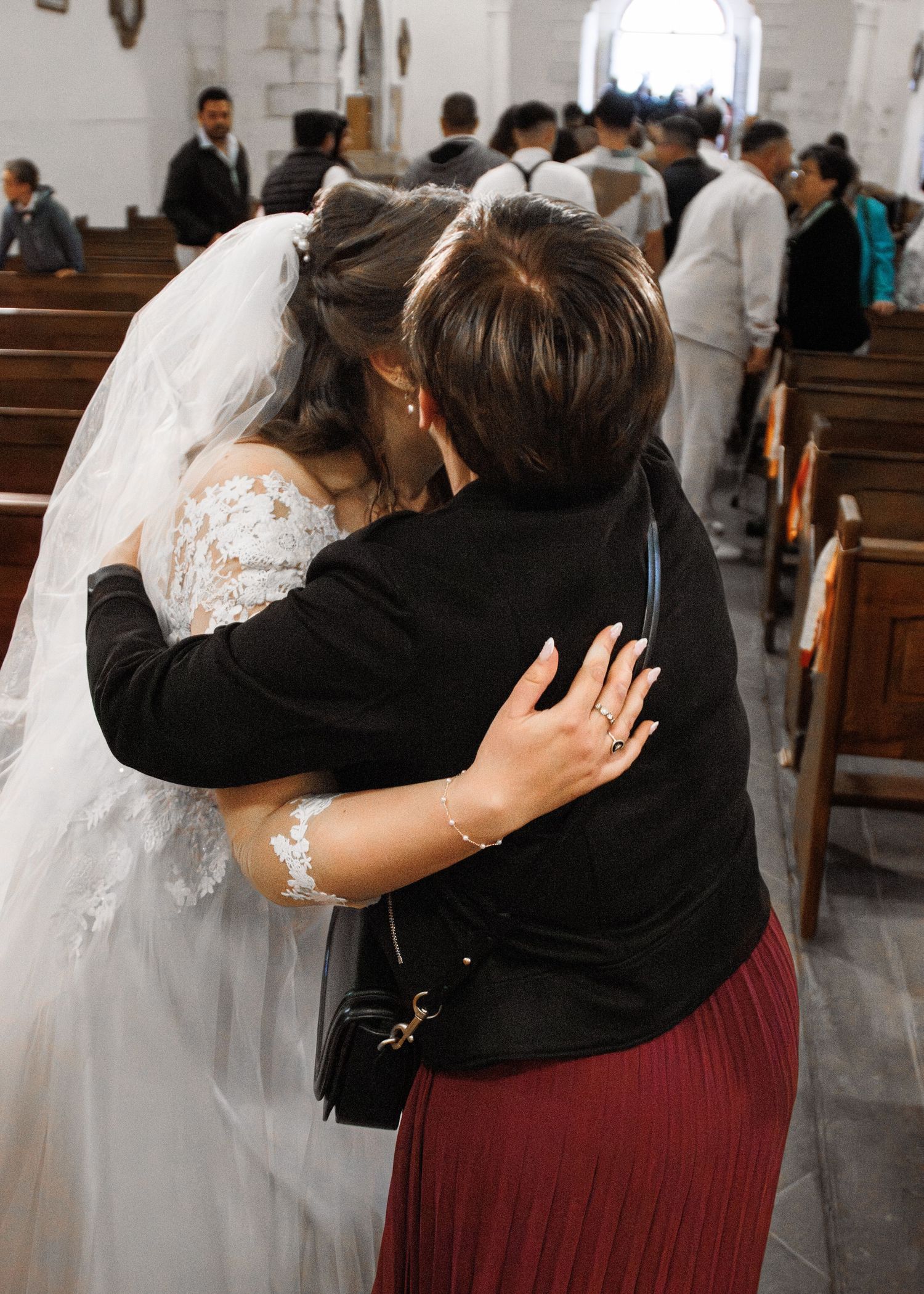 Bride in white lace gown and veil shares an emotional hug with a guest in a church during wedding ceremony.