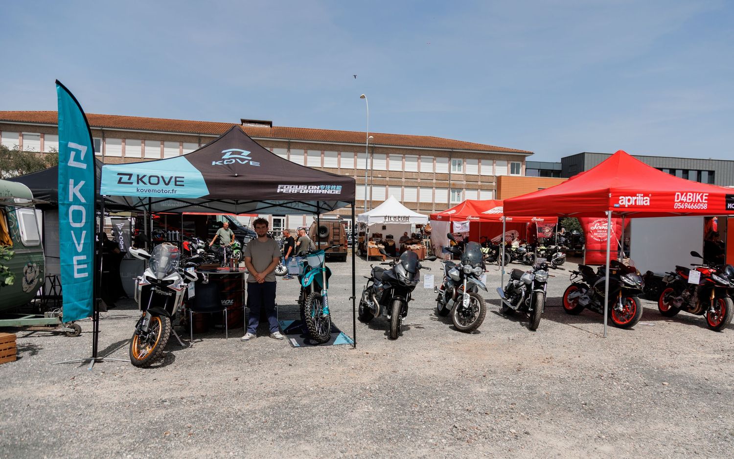 Outdoor motorcycle expo with Kove and Aprilia branded tents displaying various bikes on a gravel lot near a large building.