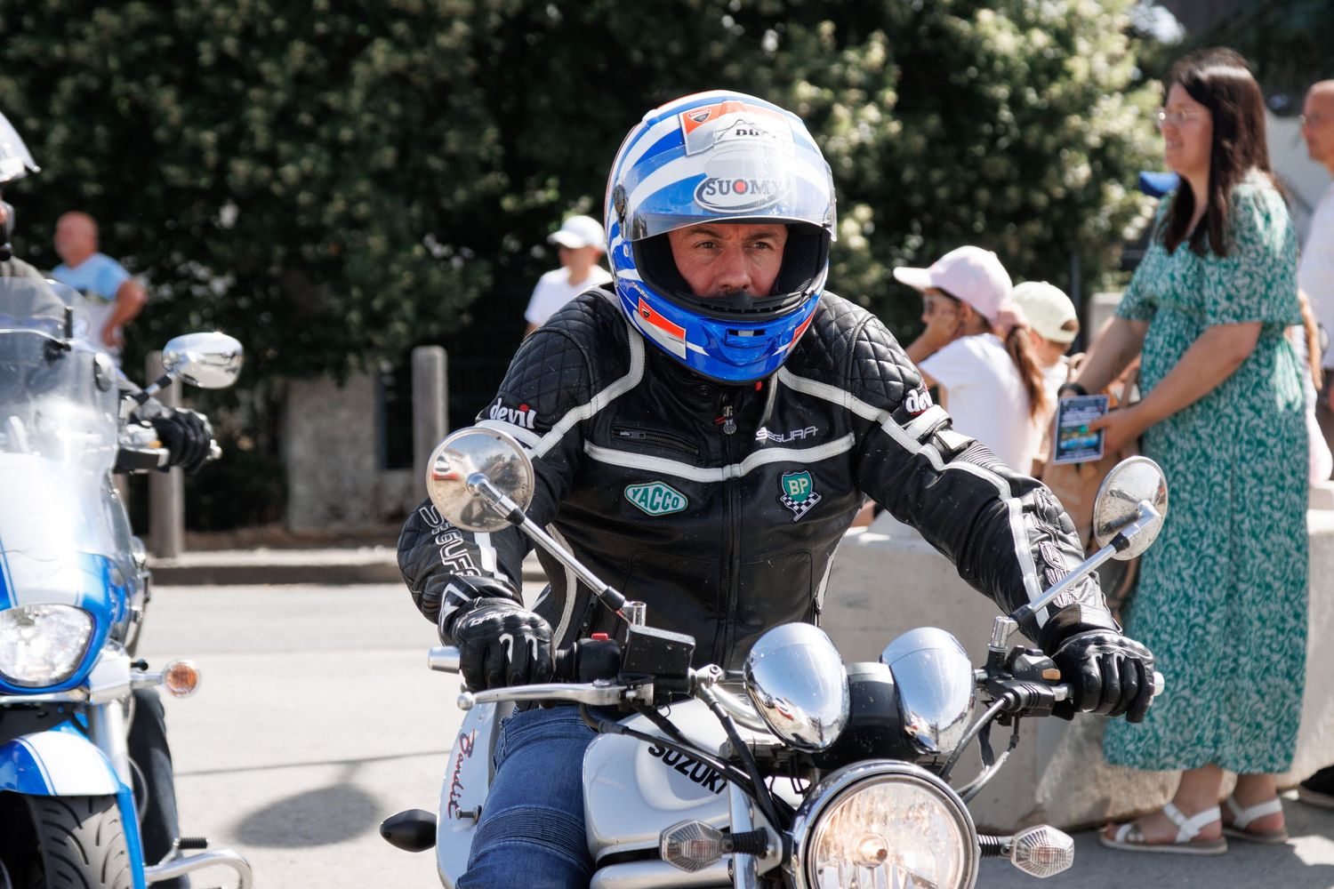 Motorcyclist wearing a blue helmet and leather jacket rides through a sunny outdoor event with spectators in background.