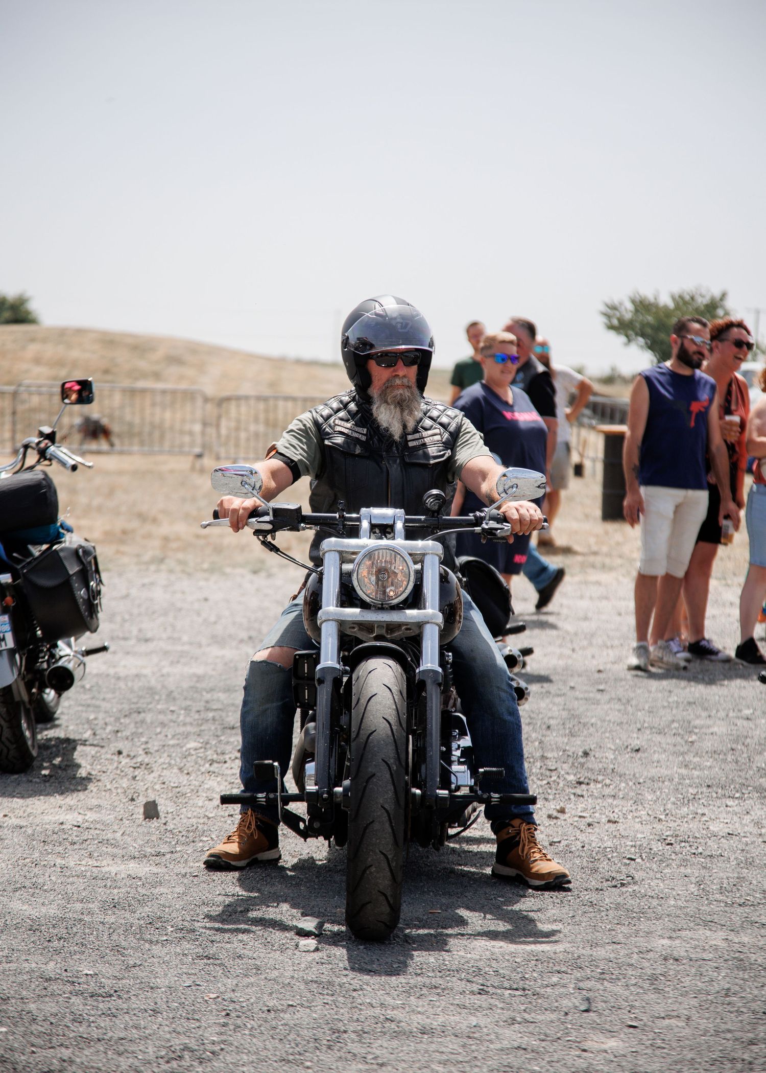 Bearded man in helmet riding a Harley-Davidson motorcycle on gravel with spectators watching in the background.