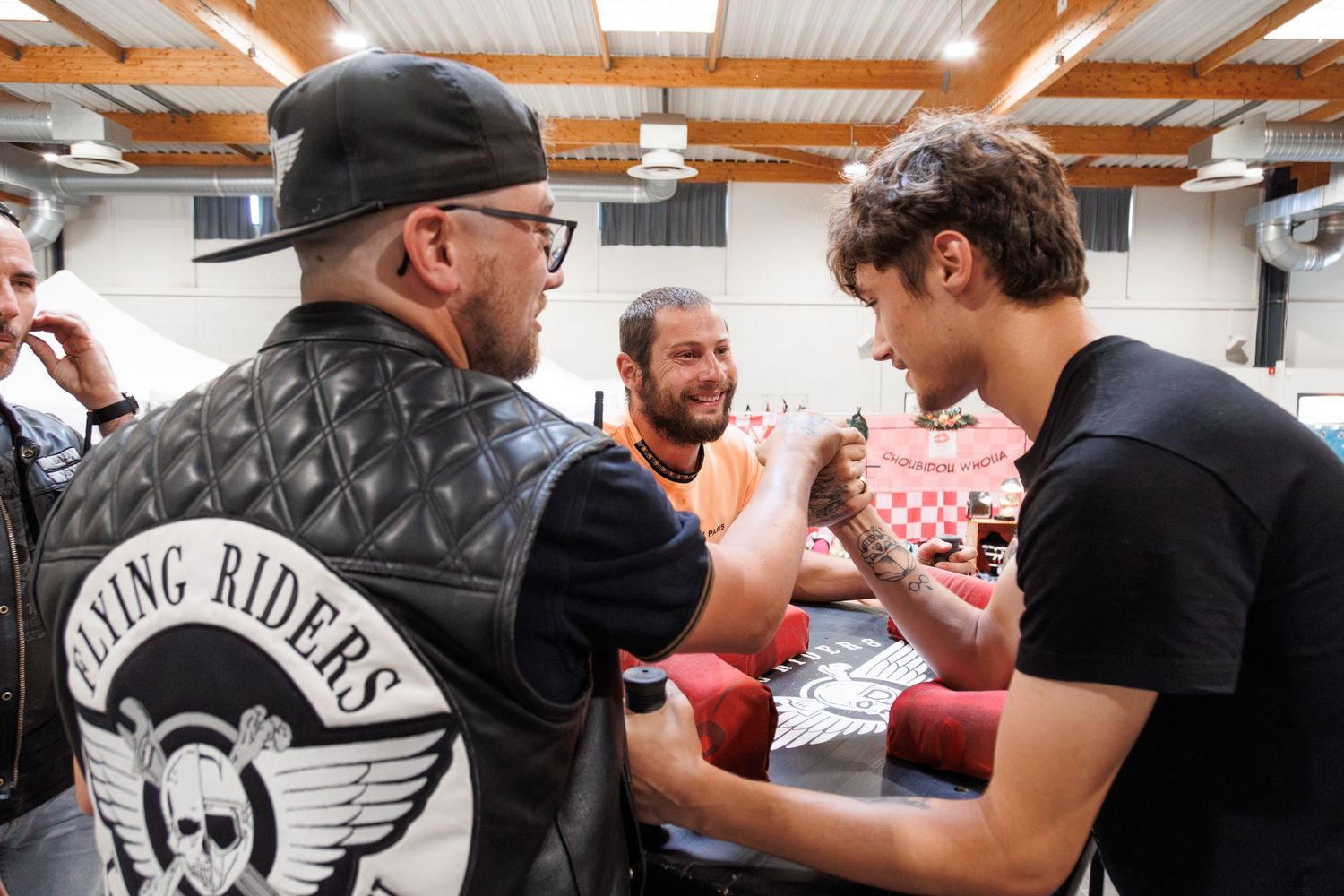 Three men chat at an indoor event, one wearing a Flying Riders biker jacket with skull logo.