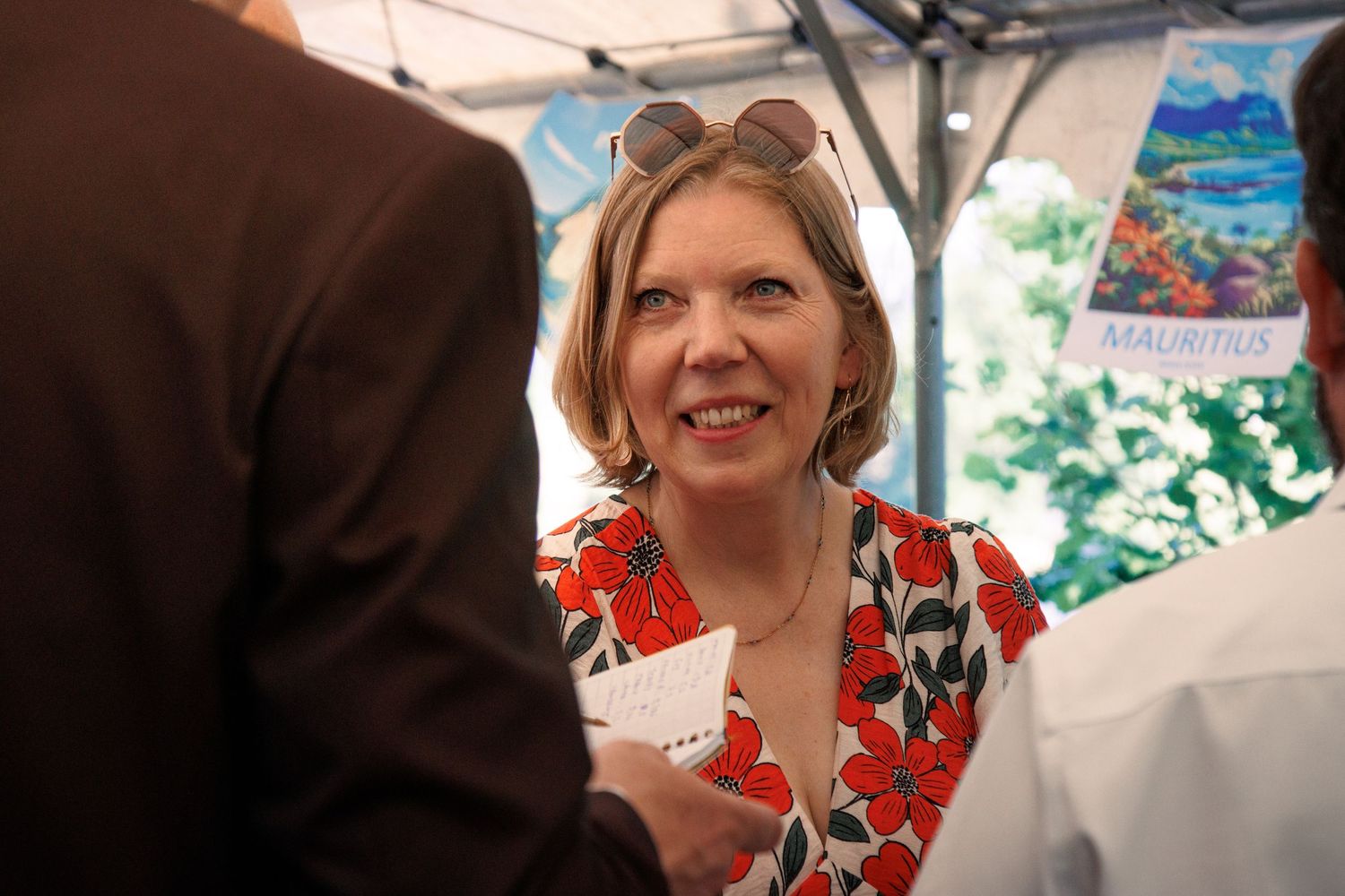 Smiling blonde woman in red floral dress with sunglasses on head at wedding reception event.