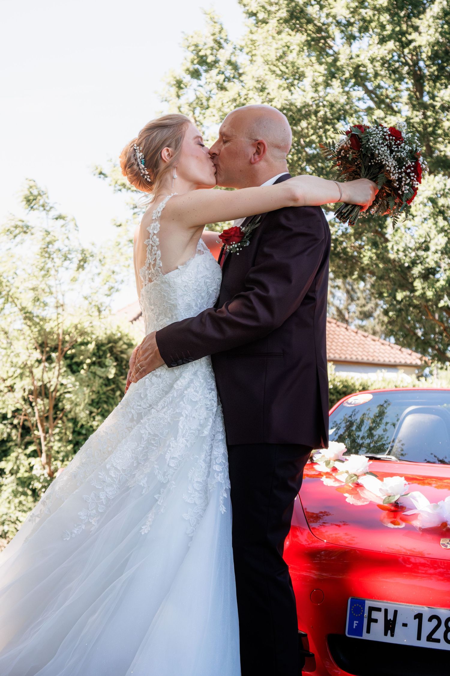 Bride and groom sharing a romantic kiss beside a decorated red car on their wedding day.