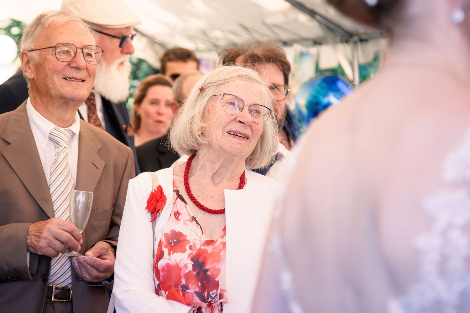 Elderly couple in formal attire smiling warmly at bride during outdoor wedding reception under tent.